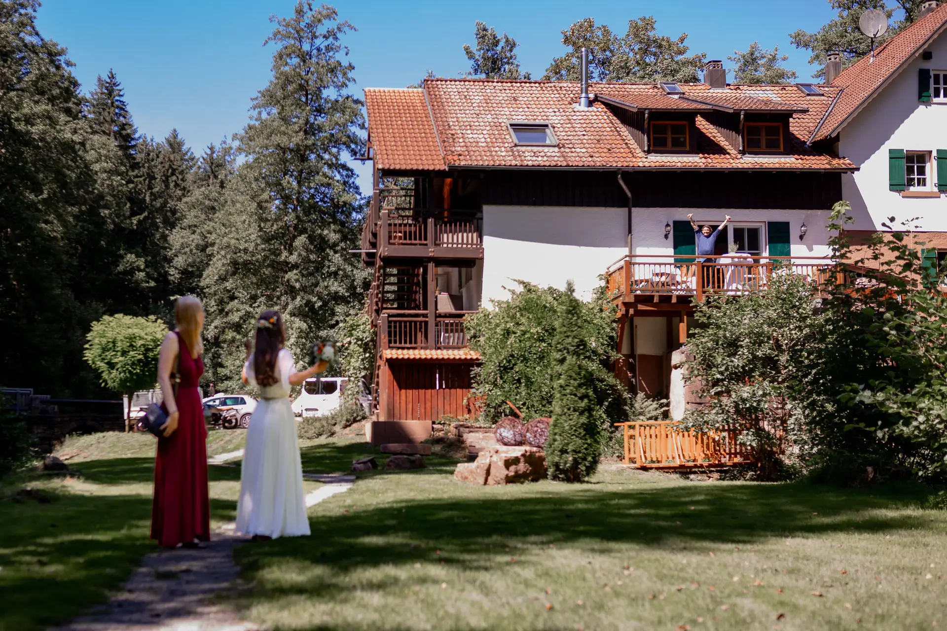 20230811 14 32 24 Hochzeit Elztal Hochzeitsfotograf Heiraten Heidelberg Landgasthof Heidersbacher Muehle 4K8A0897.DK9k5kcB Braut blickt bewundernd auf das rote Holzhaus mit Balkon und Blumendekoration der Heidersbacher Mühle