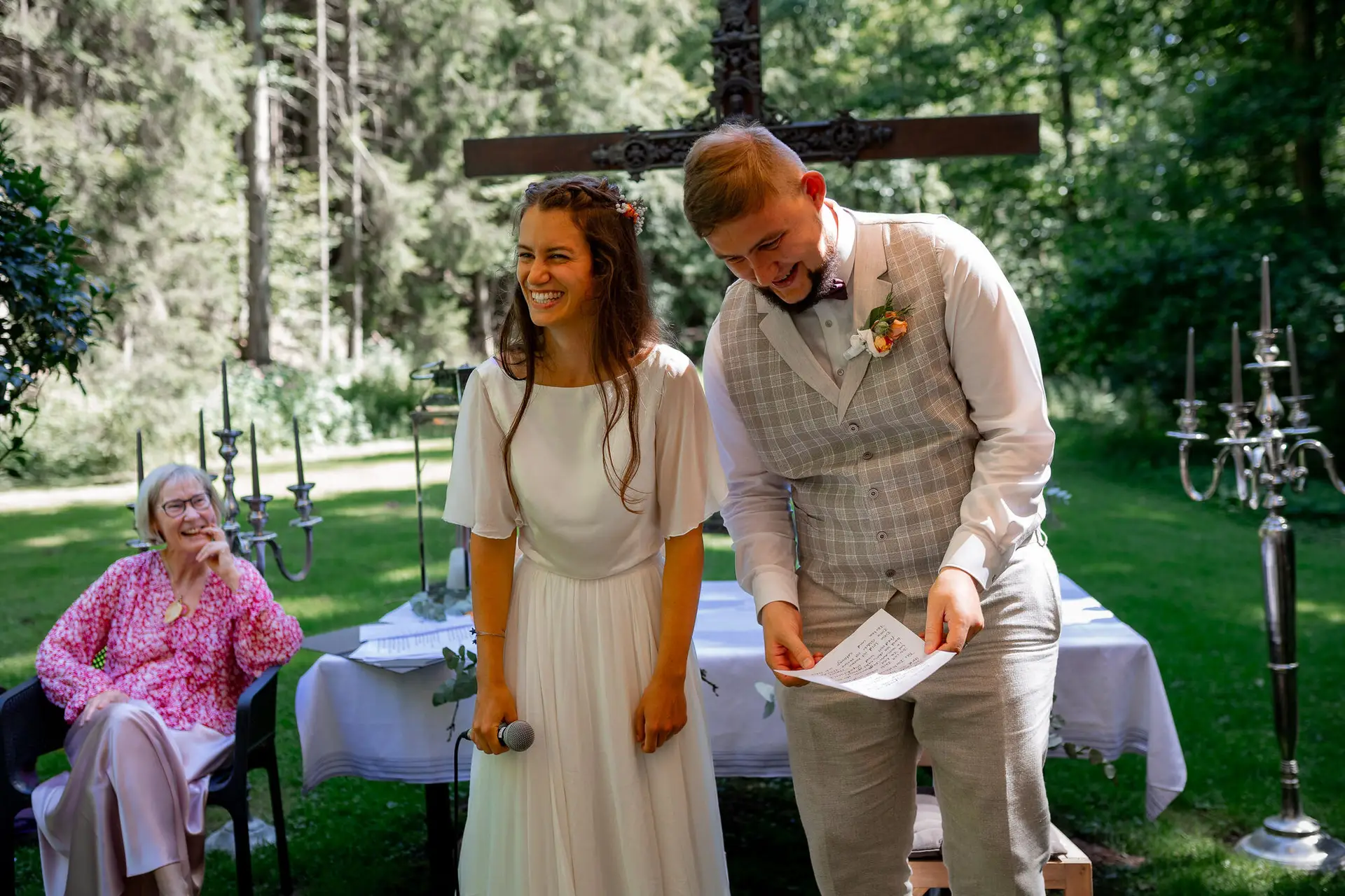 20230811 14 57 51 Hochzeit Elztal Hochzeitsfotograf Heiraten Heidelberg Landgasthof Heidersbacher Muehle 4K8A1538.Bny16aFx Brautpaar steht gemeinsam vor dem Holzkreuz auf der Wiese, Traurednerin lächelt im Hintergrund