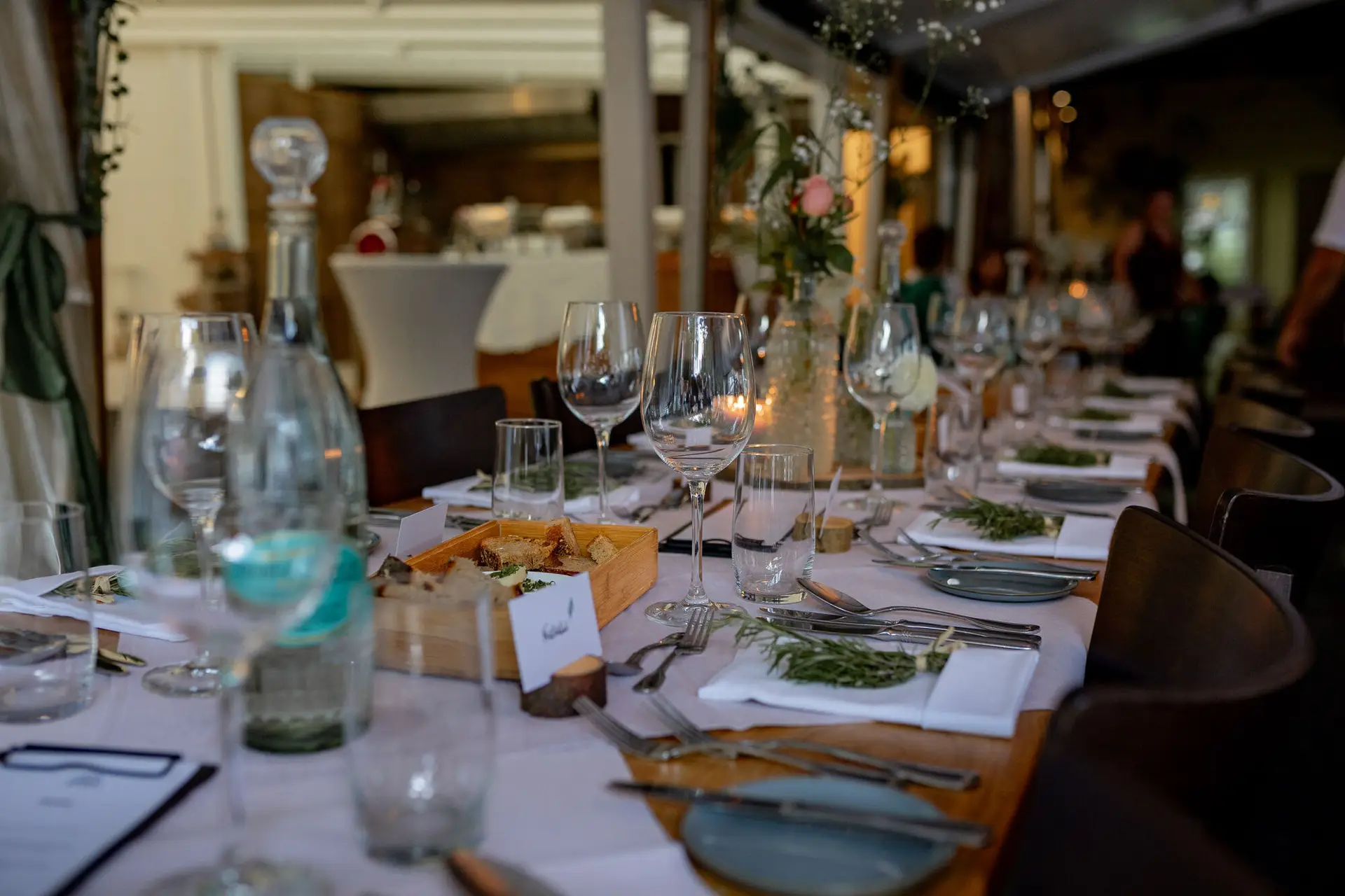 20230811 18 39 28 Hochzeit Elztal Hochzeitsfotograf Heiraten Heidelberg Landgasthof Heidersbacher Muehle 4K8A7238.CYYqIf4E Romantisch gedeckter Abendtisch mit Blumenarrangements und Kerzenlicht, Gäste im Hintergrund
