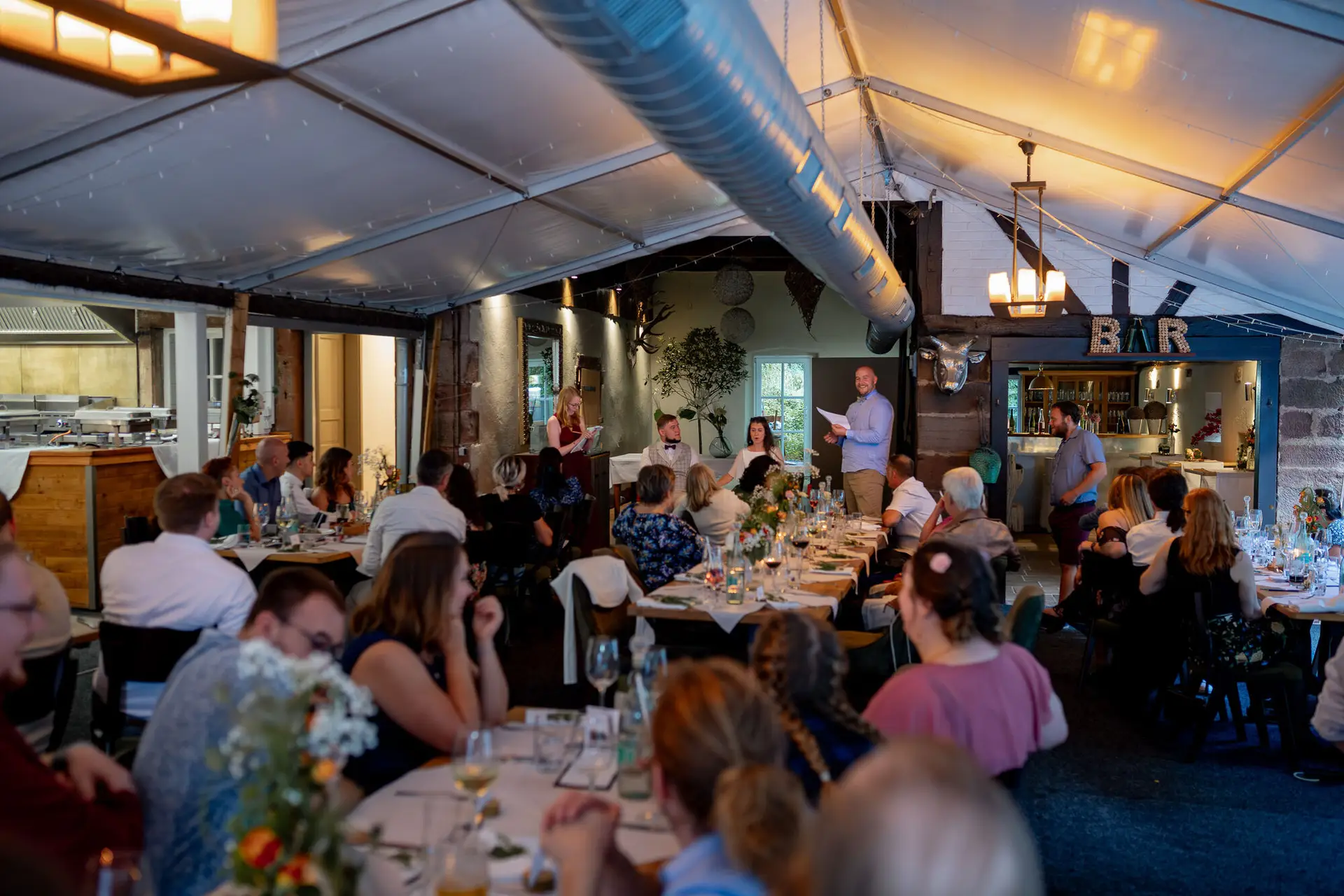 20230811 20 19 22 Hochzeit Elztal Hochzeitsfotograf Heiraten Heidelberg Landgasthof Heidersbacher Muehle 4K8A7431 Verbessert RR.BoGxWK9z Große Hochzeitsgesellschaft beim Abendessen, Redner mit Mikrofon hält Ansprache an den Tischen