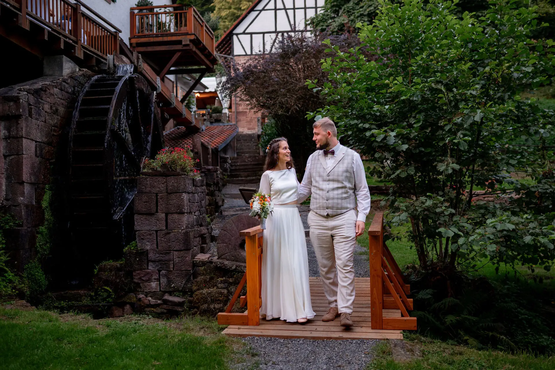 20230811 20 41 27 Hochzeit Elztal Hochzeitsfotograf Heiraten Heidelberg Landgasthof Heidersbacher Muehle 4K8A7851.B6kW91ta Brautpaar steht auf der kleinen Holzbrücke über den malerischen Wasserfall der Heidersbacher Mühle