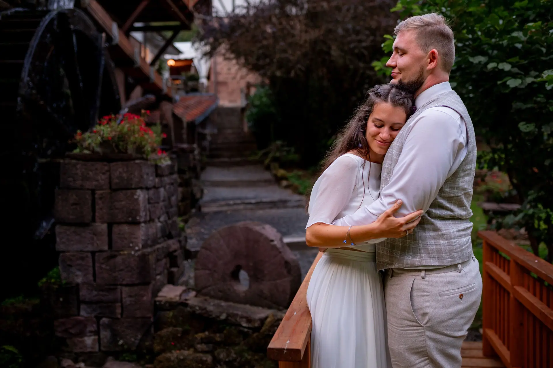 20230811 20 43 09 Hochzeit Elztal Hochzeitsfotograf Heiraten Heidelberg Landgasthof Heidersbacher Muehle 4K8A7942.Dwizx5SY Brautpaar umarmt sich zärtlich beim alten Wasserrad im abendlichen Mühlengarten