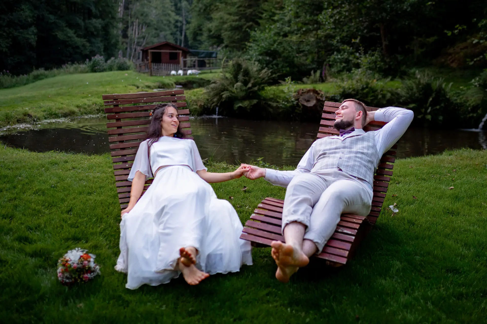 20230811 21 05 06 Hochzeit Elztal Hochzeitsfotograf Heiraten Heidelberg Landgasthof Heidersbacher Muehle 4K8A8965 Verbessert RR.CNj7R3N0 Brautpaar liegt auf Holzliegen am ruhigen Teich und hält Hände, Bäume und Natur ringsum