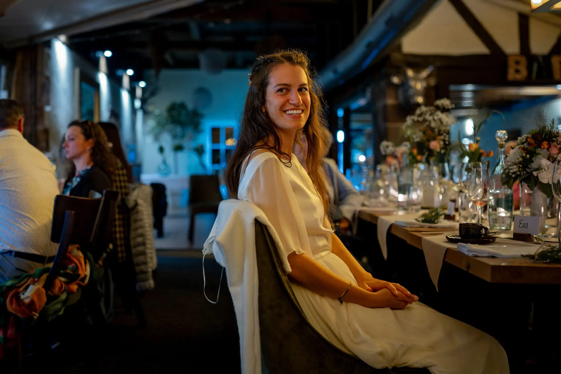 20230811 21 41 32 Hochzeit Elztal Hochzeitsfotograf Heiraten Heidelberg Landgasthof Heidersbacher Muehle 4K8A9189 Verbessert RR.BNSTdT2O Braut sitzt entspannt am Abendtisch und genießt den Moment, Gäste feiern im Hintergrund