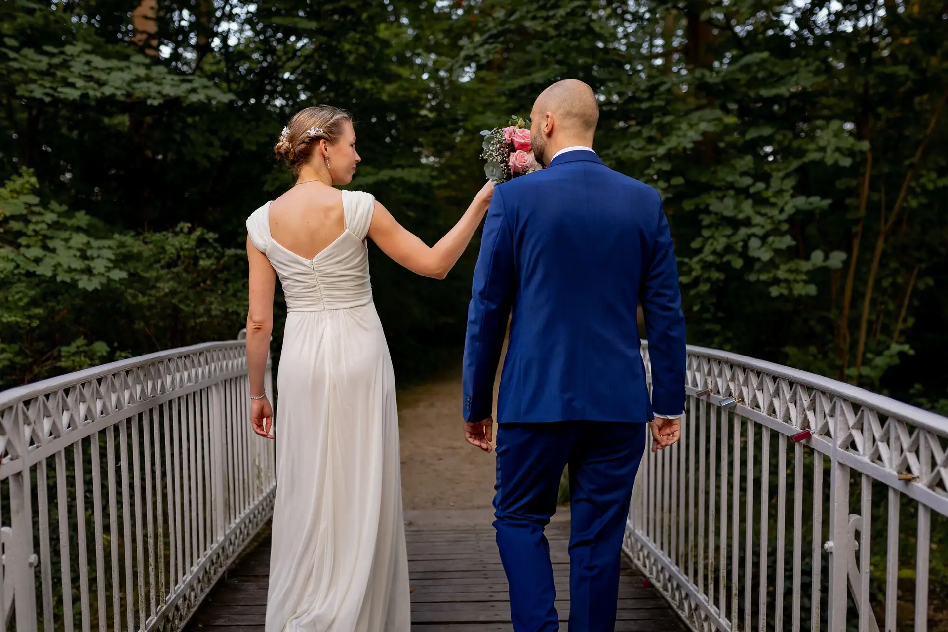 20240820 19 42 08 Schlossgarten Worms Herrnsheim 4K8A8970.B5A8DWr2 Brautpaar geht Hand in Hand über die Holzbrücke im Wald, Braut im weißen Kleid, Bräutigam im dunkelblauen Anzug