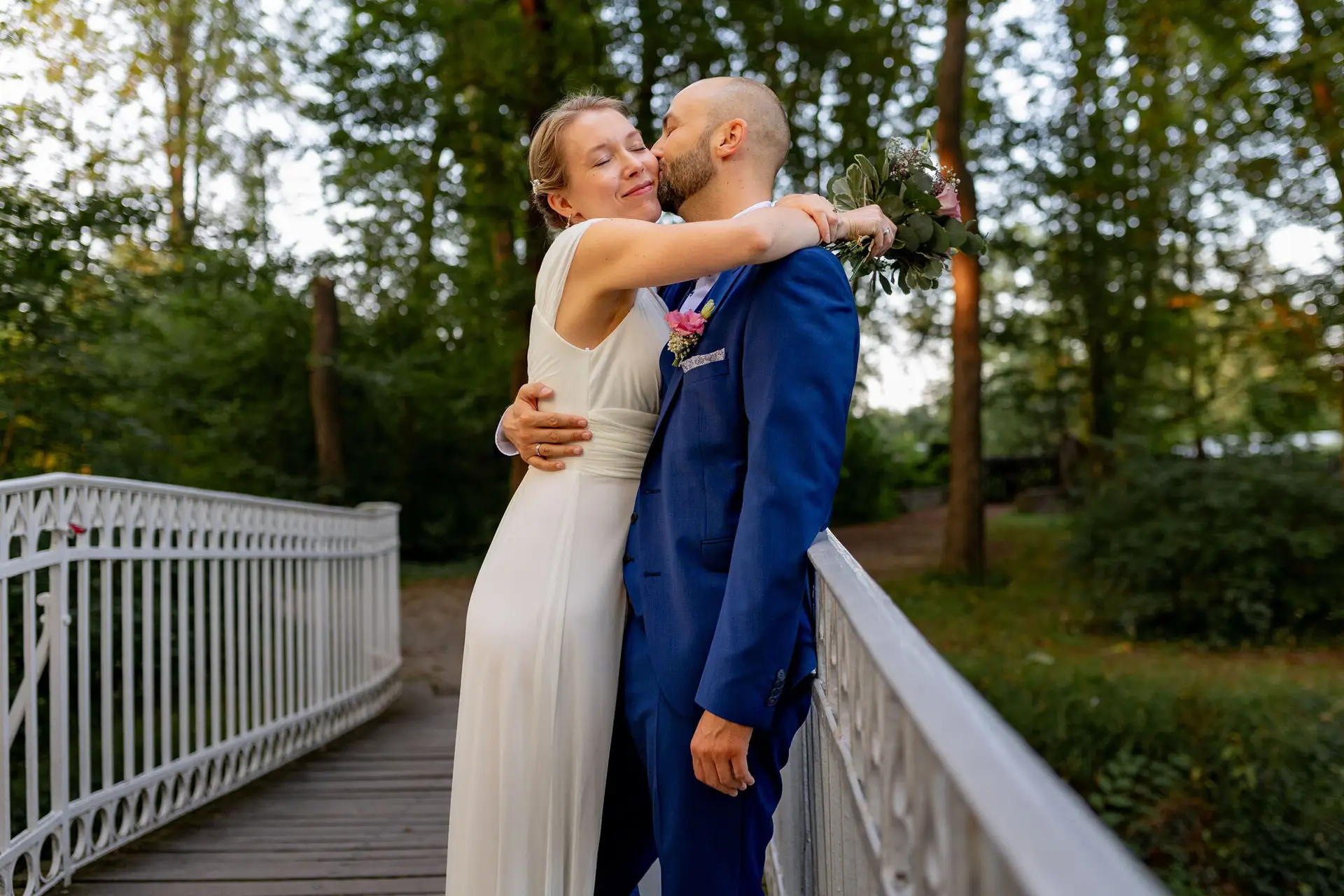 20240820 19 47 02 Schlossgarten Worms Herrnsheim 4K8A9197 Hochzeitsfotograf Matthias Butz.CjekG6DD Bräutigam umarmt Braut auf der Holzbrücke im Wald, Braut hält kleinen Blumenstrauß
