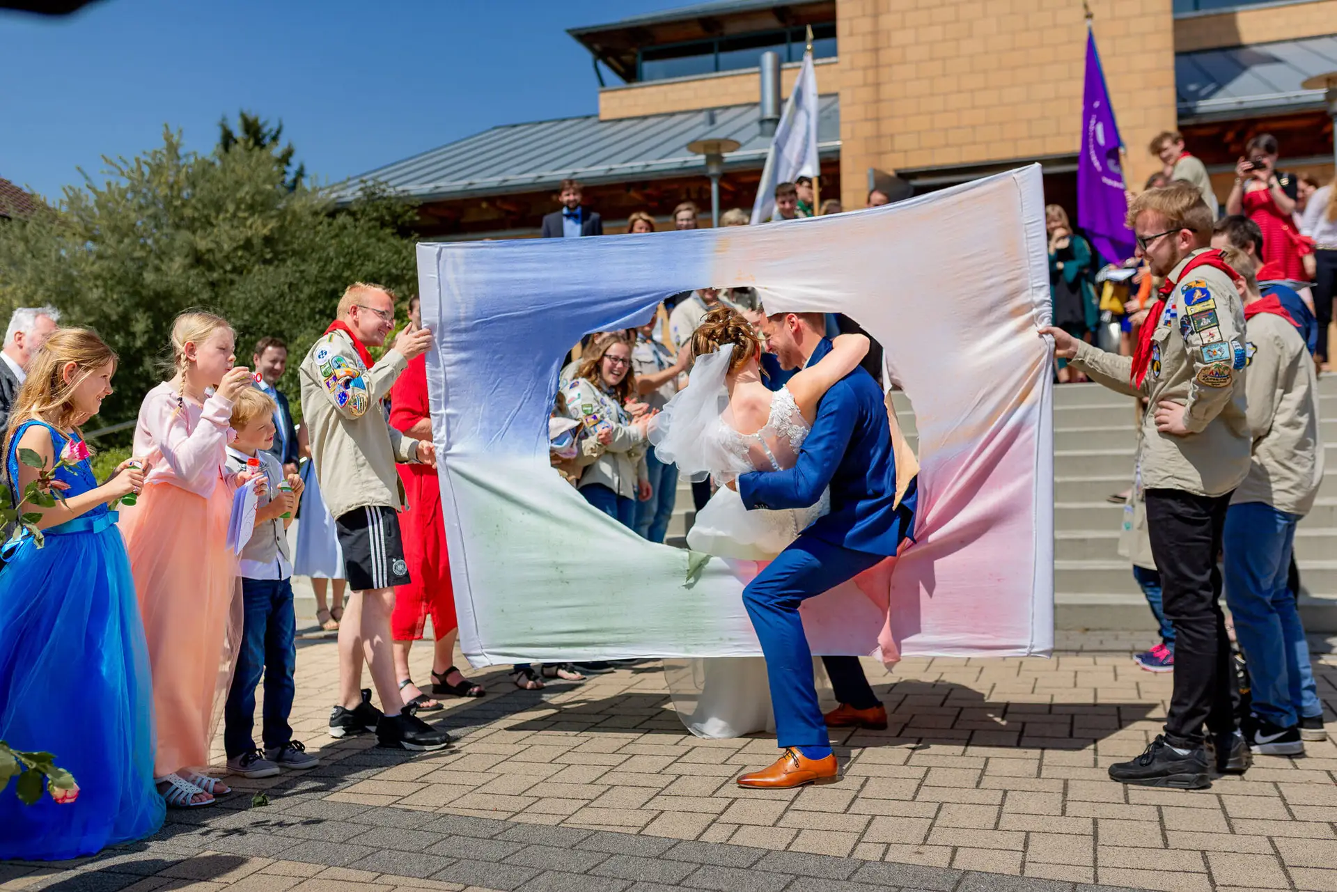 Anne Christian Hochzeitsfotograf Birkenheide Grosskarlbach Bissersheim 00017.C9PnUyO Brautpaar küsst sich leidenschaftlich unter buntem Stoffbanner vor blauem Himmel, Gäste jubeln rundherum