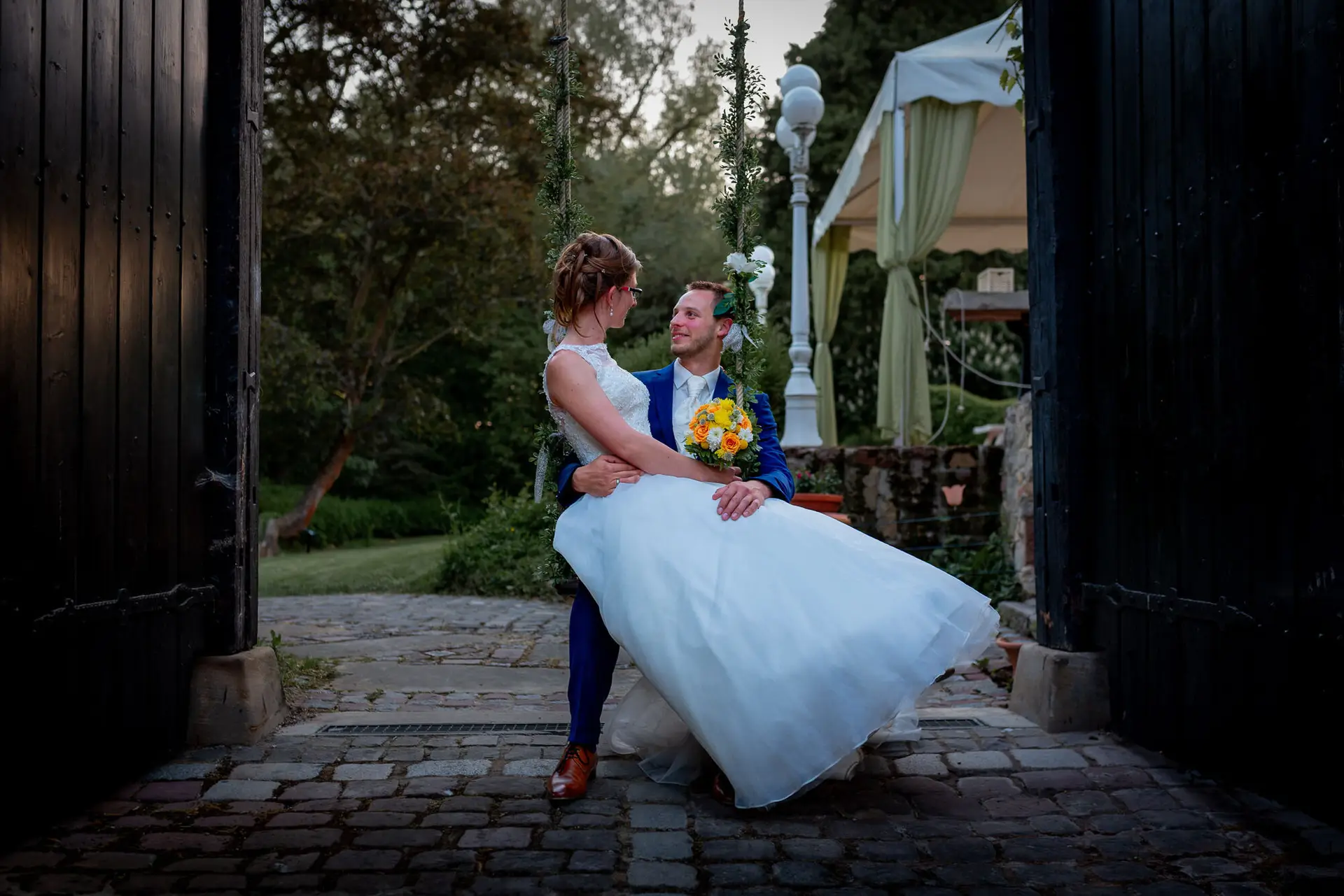 Anne Christian Hochzeitsfotograf Birkenheide Grosskarlbach Bissersheim 00040.B5NTC83p Brautpaar sitzt eng umschlungen an einem Holzzaun unter grünem Pavillon mit gelben Blüten