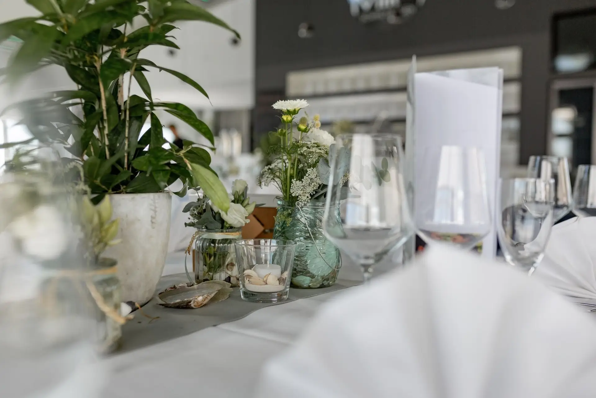 Festlich gedeckter Tisch mit weißen Blumen, Eukalyptus und Gläsern, erste Gäste kommen im Hintergrund an