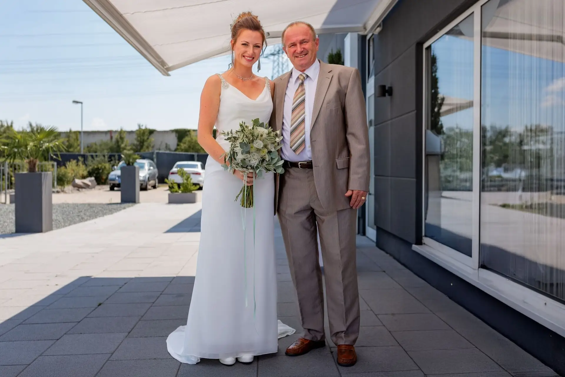 Braut im weißen Kleid mit Schleier und Blumenstrauß neben ihrem Vater im braunen Anzug vor dem Einzug