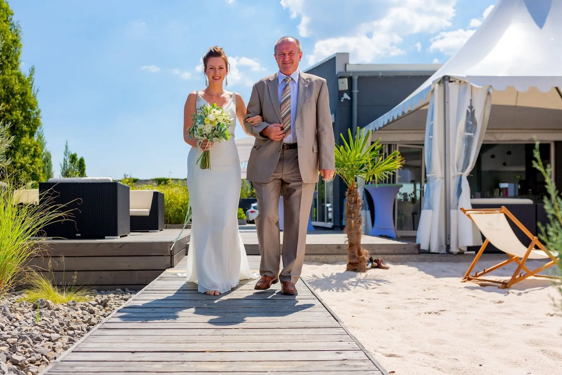 Braut wird von ihrem Vater über den Holzsteg vor dem weißen Pavillon geführt, emotionaler Einzugsmoment