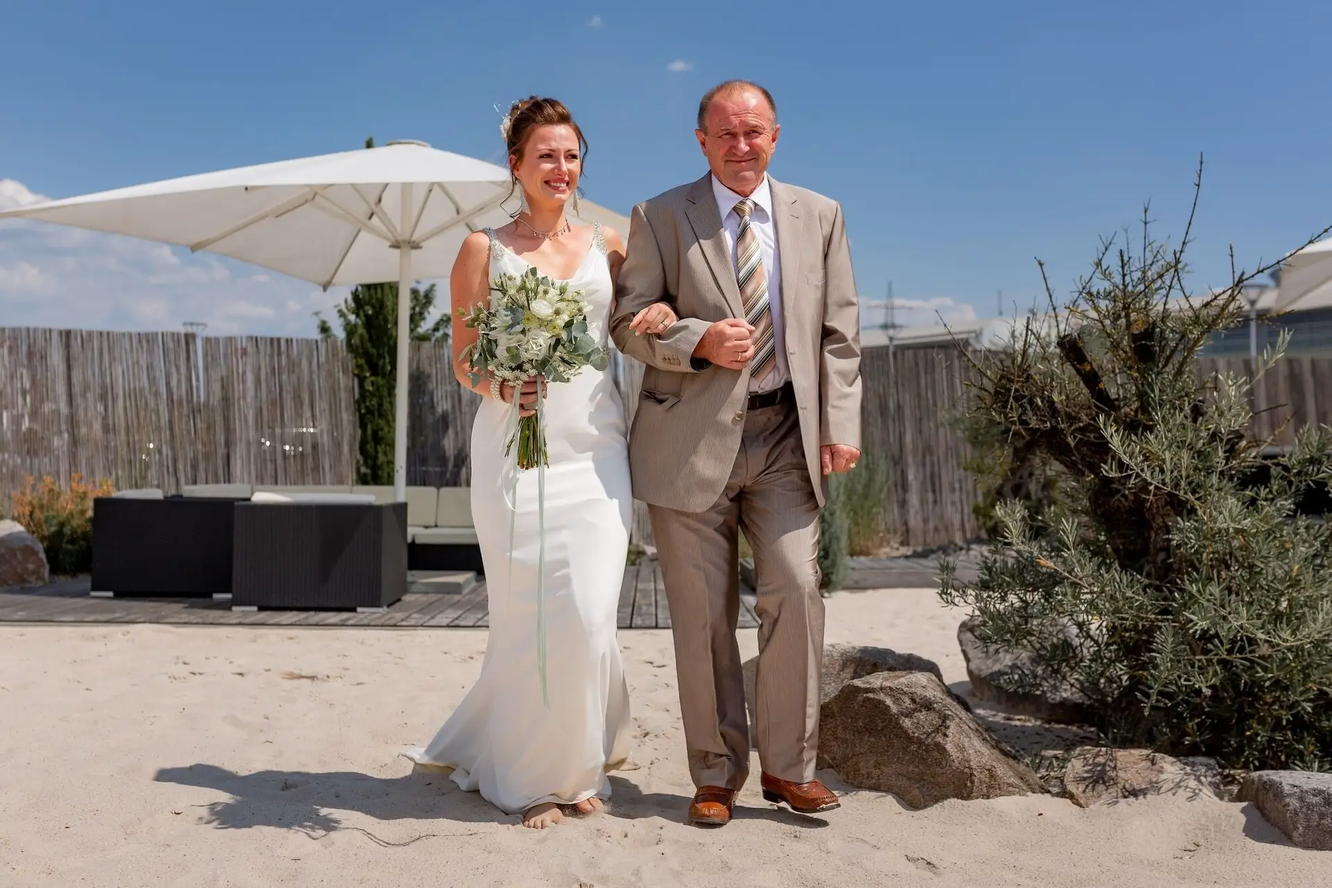 Braut schreitet mit ihrem Vater über den Sandstrand des Miio Schwetzingen zum Traualtar
