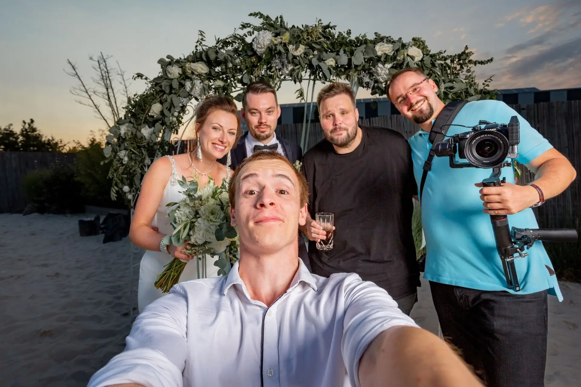 Brautpaar und Gäste machen ein fröhliches Selfie am Strand vor dem Blumenbogen