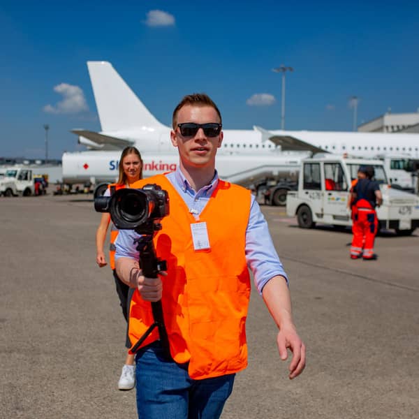 Behind the scenes: Filmer am Flughafen Köln/Bonn mit Kamera und Sicherheitsweste beim Videodreh auf dem Rollfeld vor einem Flugzeug.