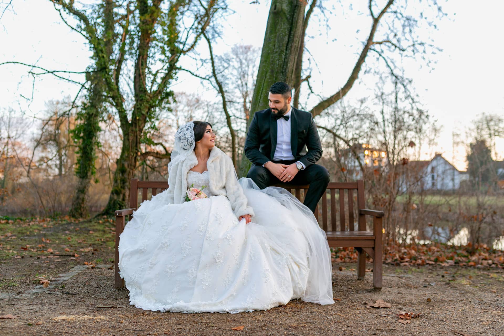 20181225 16 57 30 MK3 4545 Brautpaarshooting Worms Hochzeitsfeier Mannheim.BlqMafyp Brautpaar sitzt auf einer Holzbank im winterlichen Waldweg, Braut im weißen Fellmantel, kahle Bäume im Hintergrund