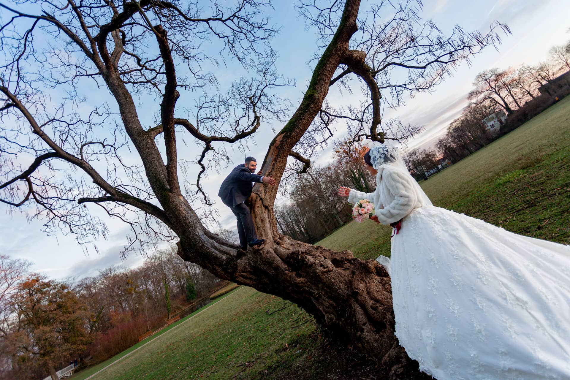 20181225 17 17 14 MK3 4661 Brautpaarshooting Worms Hochzeitsfeier Mannheim.Cigp6PxE Brautpaar sitzt und hält sich auf einem knorrigen alten Baum, historisches Haus des Schlossgartens im Hintergrund