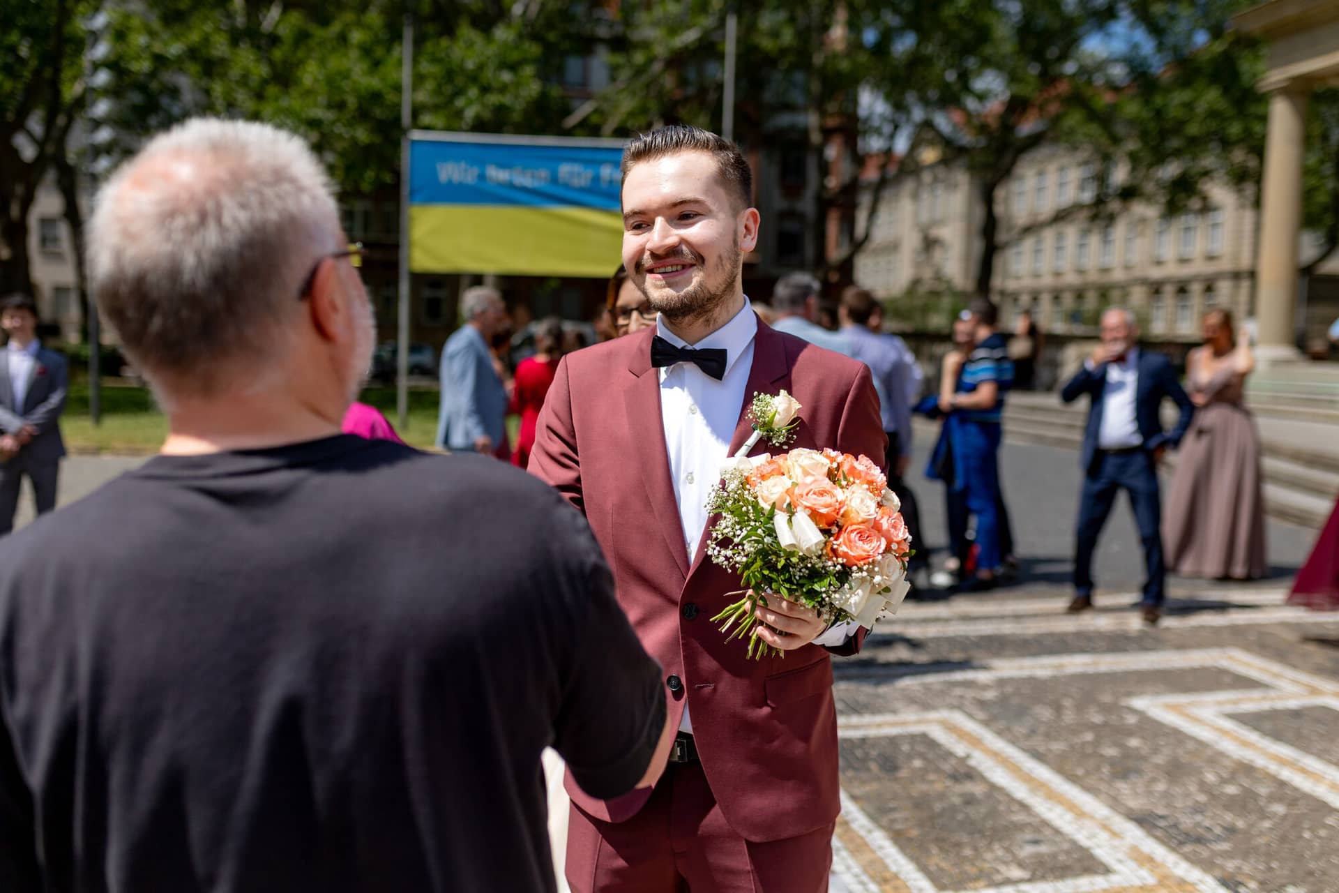 20230610 14 05 32 4K8A1143 Hochzeitsfotograf Mannheim Christuskirche Neuleiningen Burgschenke.CNzgyPPr Bräutigam überreicht einen roten Blumenstrauß vor dem belebten Stadtbild Mannheims nach der Trauung