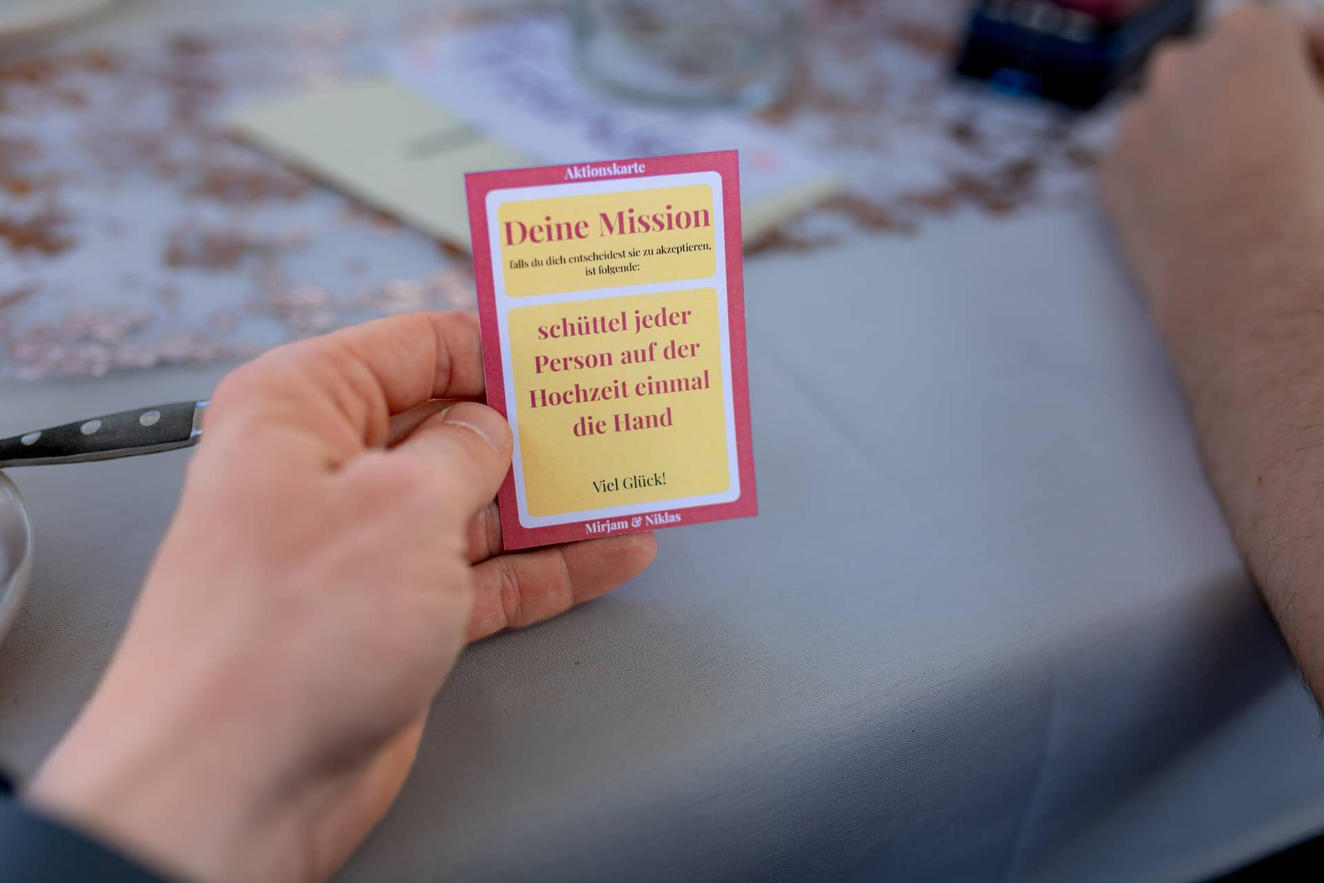20230610 17 03 20 4K8A2517 Hochzeitsfotograf Mannheim Christuskirche Neuleiningen Burgschenke.BUP10SIB Hand hält eine rote Hochzeitskarte mit der Aufschrift „Viel Glück! Mirjam & Niklas" vor verschwommenem Hintergrund