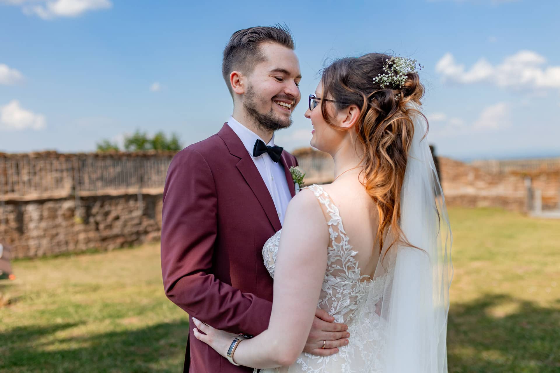 20230610 17 51 54 4K8A3480 Hochzeitsfotograf Mannheim Christuskirche Neuleiningen Burgschenke.CJ09dshA Brautpaar lächelt sich glücklich vor dem mittelalterlichen Steinwall der Burg Neuleiningen bei blauem Himmel an