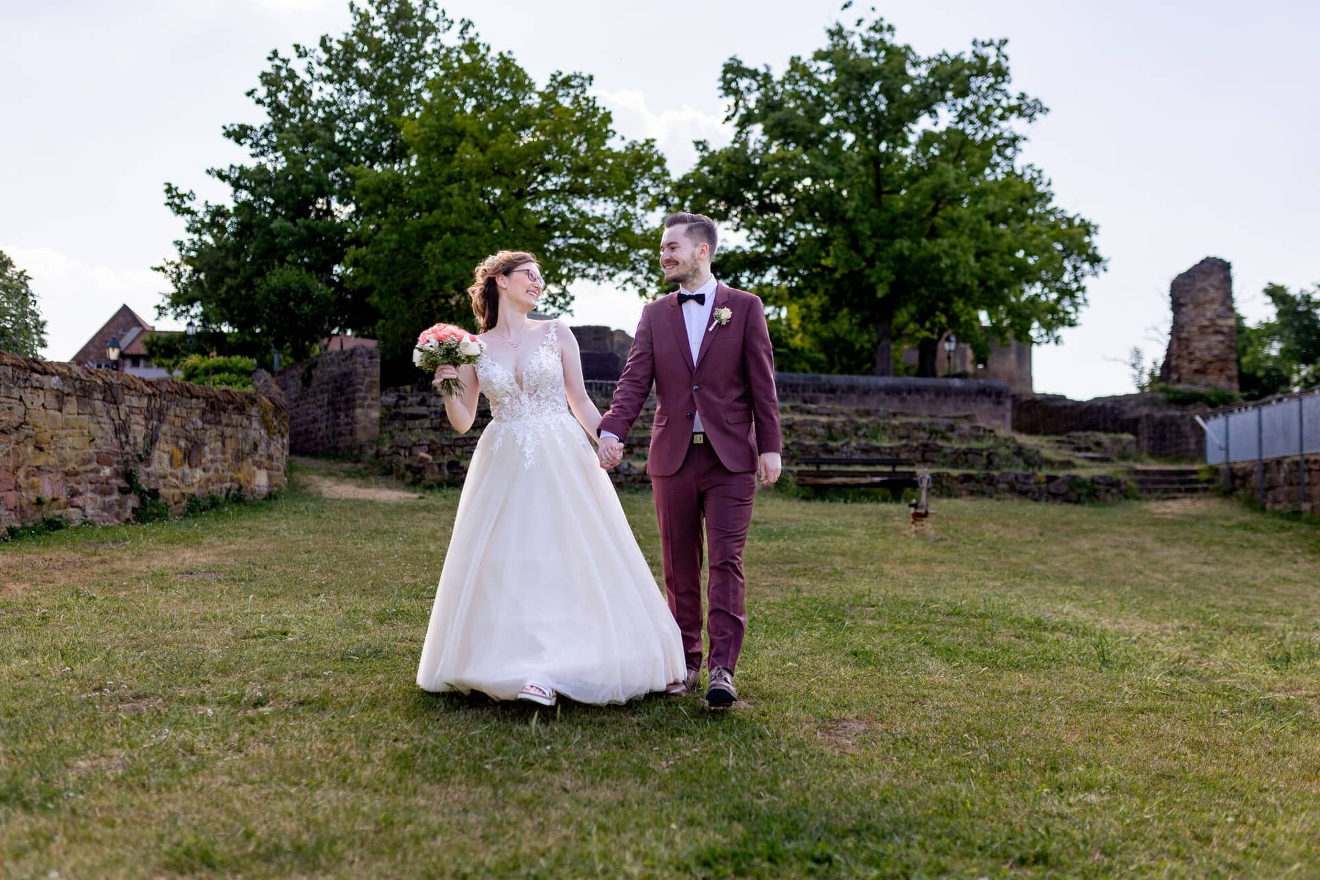 20230610 17 55 11 4K8A3786 Hochzeitsfotograf Mannheim Christuskirche Neuleiningen Burgschenke.C RkIf7M Brautpaar hält Händchen und spaziert auf dem Rasen entlang des historischen Steinwalls der Burganlage