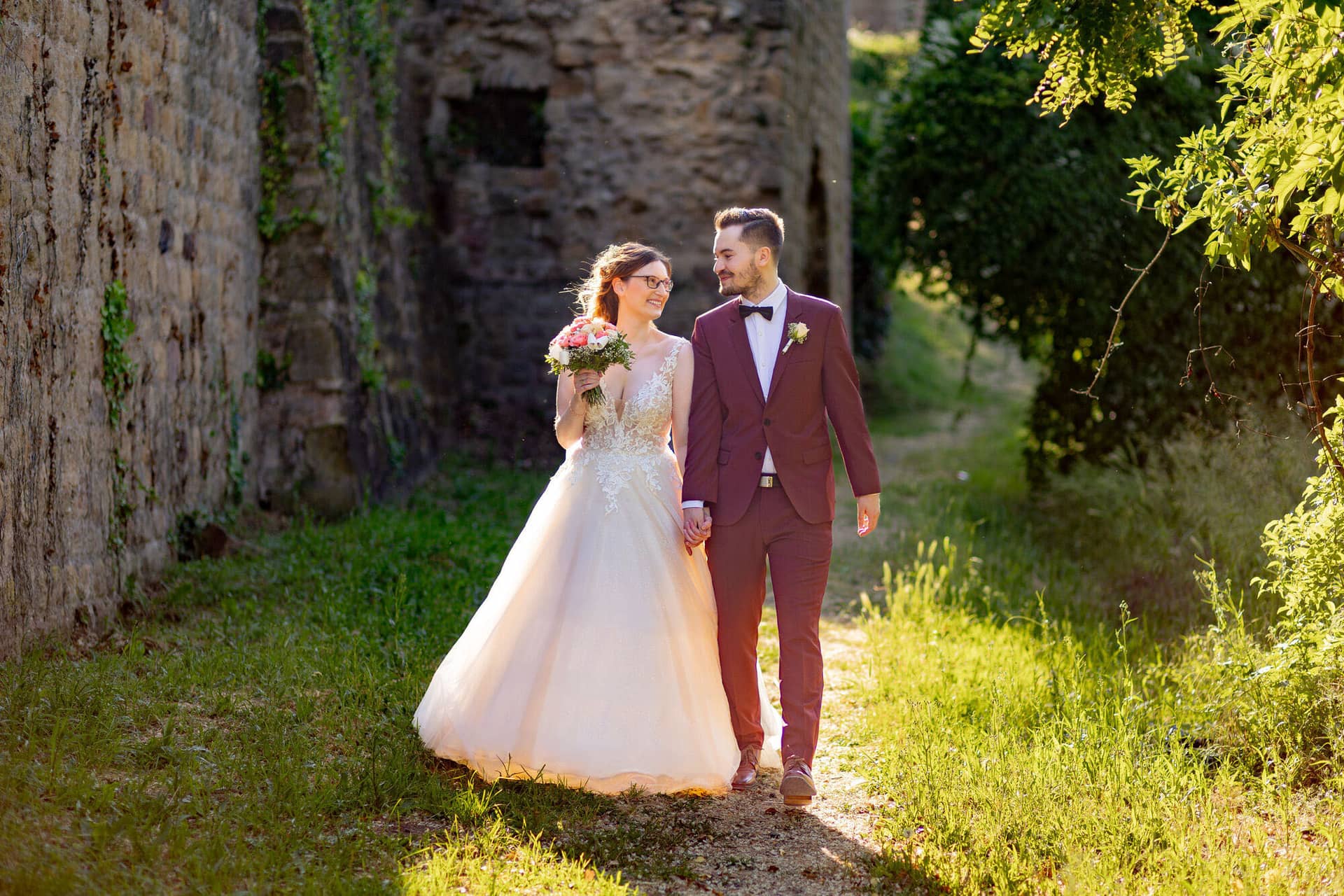 20230610 18 16 22 4K8A4585 Hochzeitsfotograf Mannheim Christuskirche Neuleiningen Burgschenke.94htwBNi Brautpaar hält Hände auf dem Pfad neben dem alten Steinkeller im goldenen Abendlicht der Burg Neuleiningen