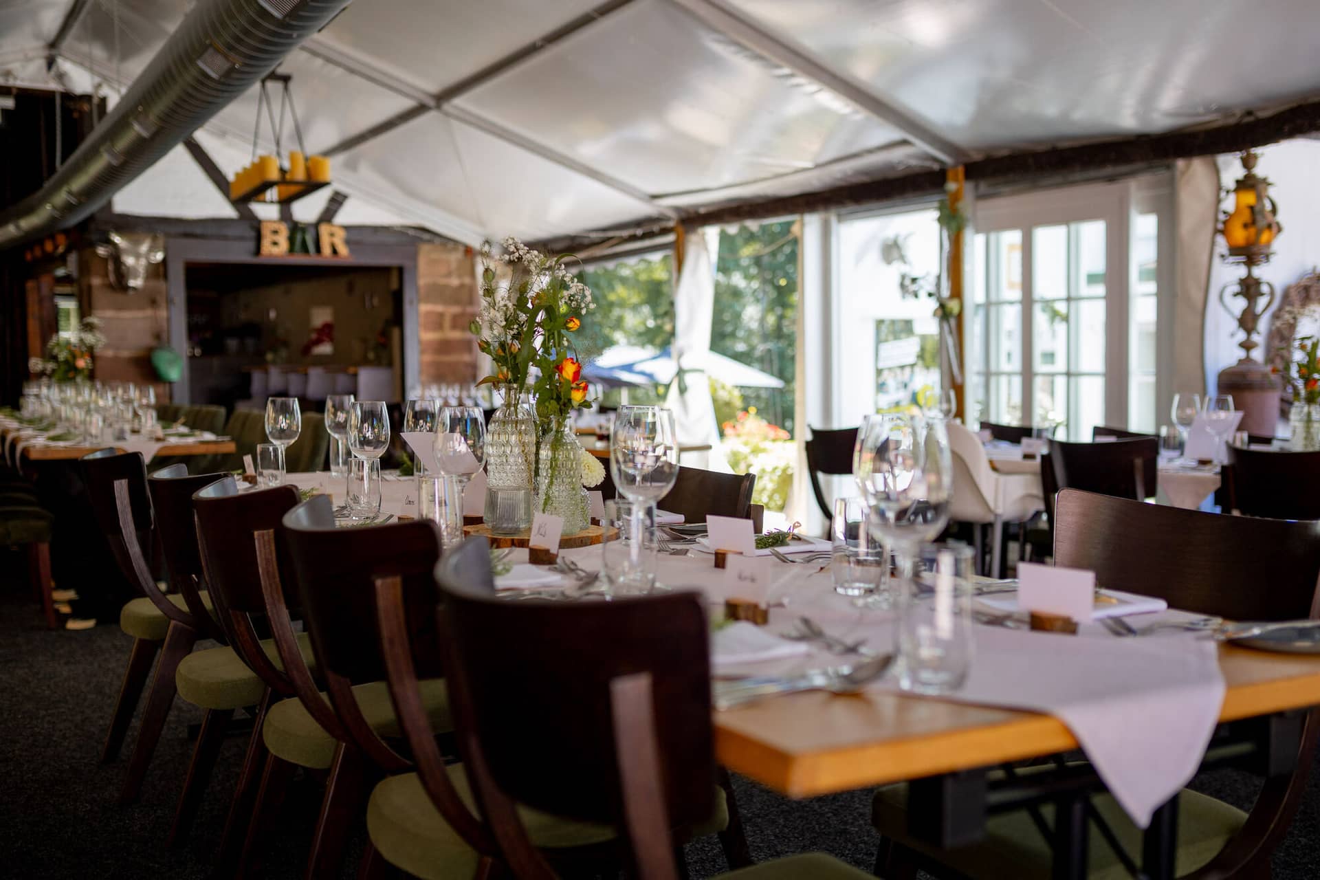 20230811 13 26 47 Hochzeit Elztal Hochzeitsfotograf Heiraten Heidelberg Landgasthof Heidersbacher Muehle 4K8A0346.DIZUQmxf Weißes Hochzeitszelt mit elegant gedeckten Tischen, grünen Sesseln und rustikaler Bar mit Vintage-Leuchten an der Heidersbacher Mühle