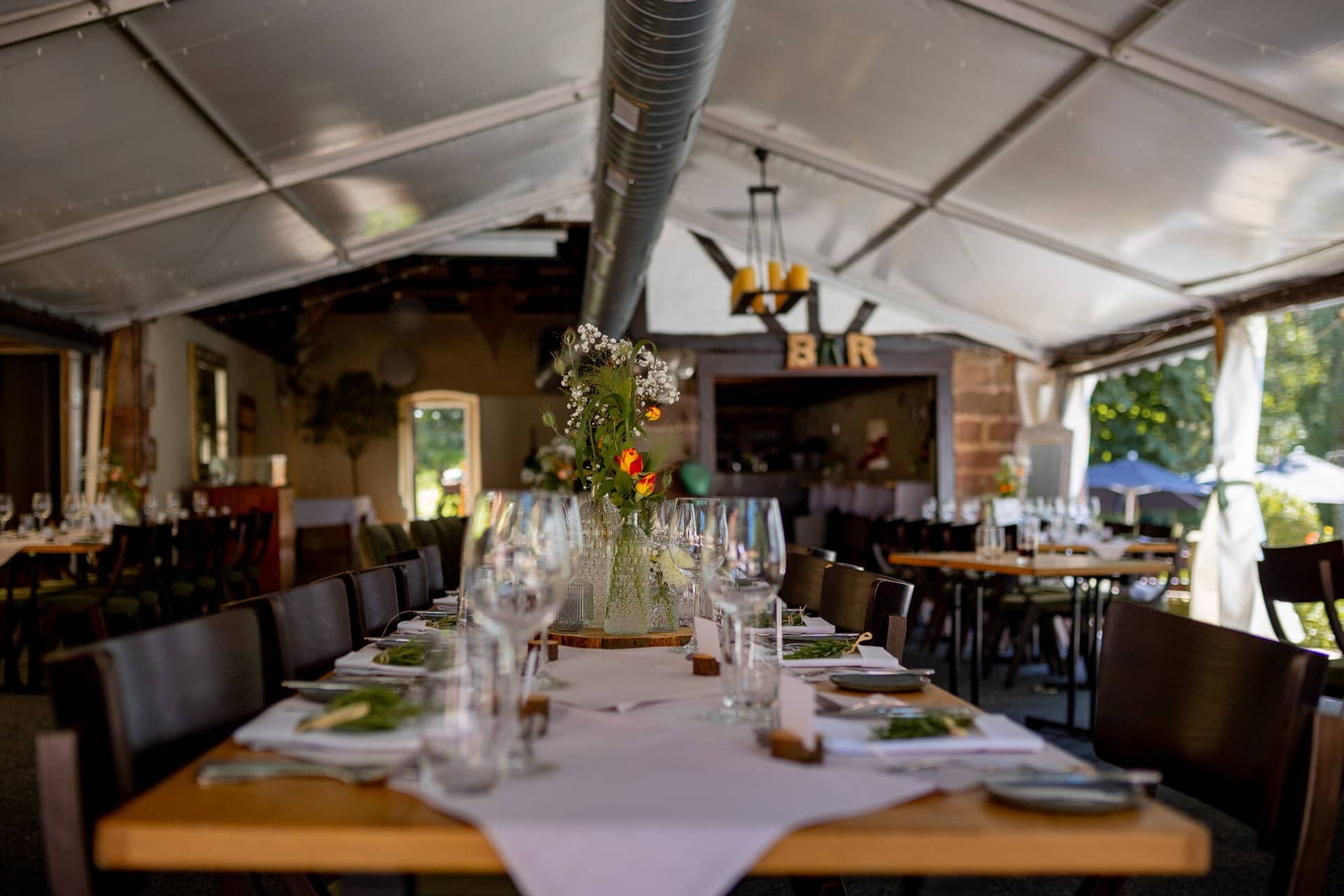 20230811 13 26 58 Hochzeit Elztal Hochzeitsfotograf Heiraten Heidelberg Landgasthof Heidersbacher Muehle 4K8A0352.DWX AhPy Langer Holztisch im hellen Zelt mit weißer Blumenvase und Bar-Schild der Location