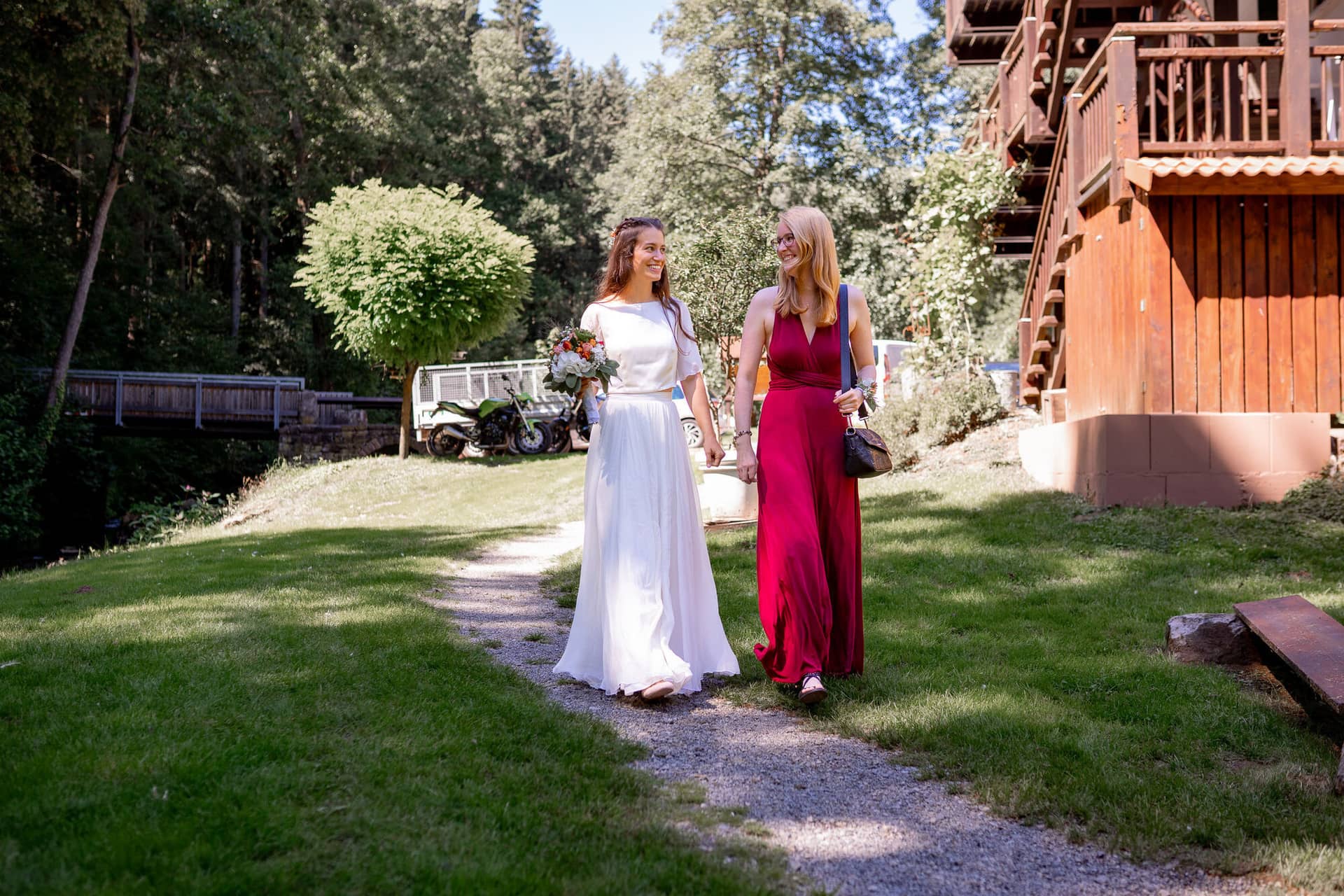 20230811 14 31 58 Hochzeit Elztal Hochzeitsfotograf Heiraten Heidelberg Landgasthof Heidersbacher Muehle 4K8A0857.BMLR4k A Braut und Brautjungfer in Rot spazieren auf dem Feldweg durch den Elztal-Wald, altes Holzhaus im Hintergrund
