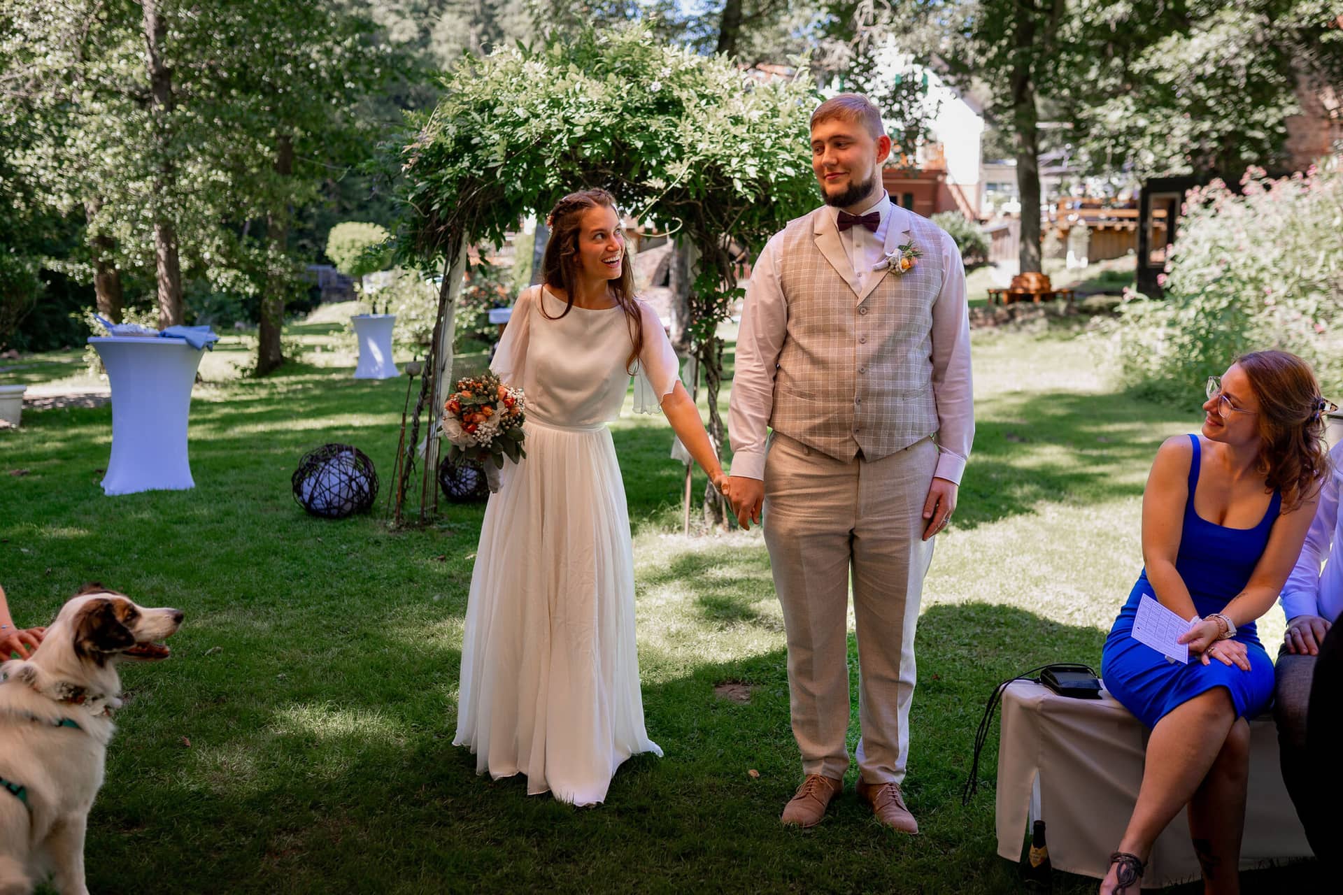 20230811 14 33 26 Hochzeit Elztal Hochzeitsfotograf Heiraten Heidelberg Landgasthof Heidersbacher Muehle 4K8A1035.CeJSMcvU Brautpaar hält sich die Hände im Garten bei Beginn der Zeremonie, Familienhund sitzt ruhig dabei