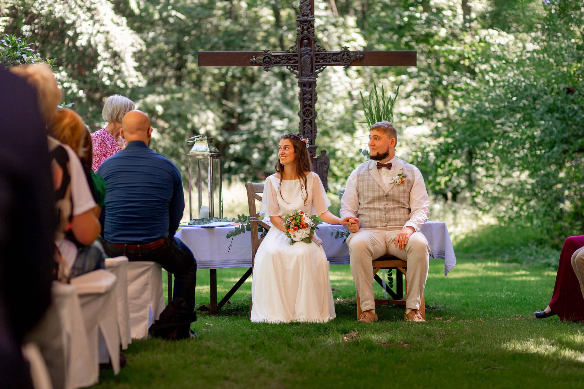 20230811 14 37 00 Hochzeit Elztal Hochzeitsfotograf Heiraten Heidelberg Landgasthof Heidersbacher Muehle MK3 8839.BYCTRRuJ Brautpaar sitzt gemeinsam bei der Zeremonie vor dem Holzkreuz, Gäste umgeben sie im Halbkreis