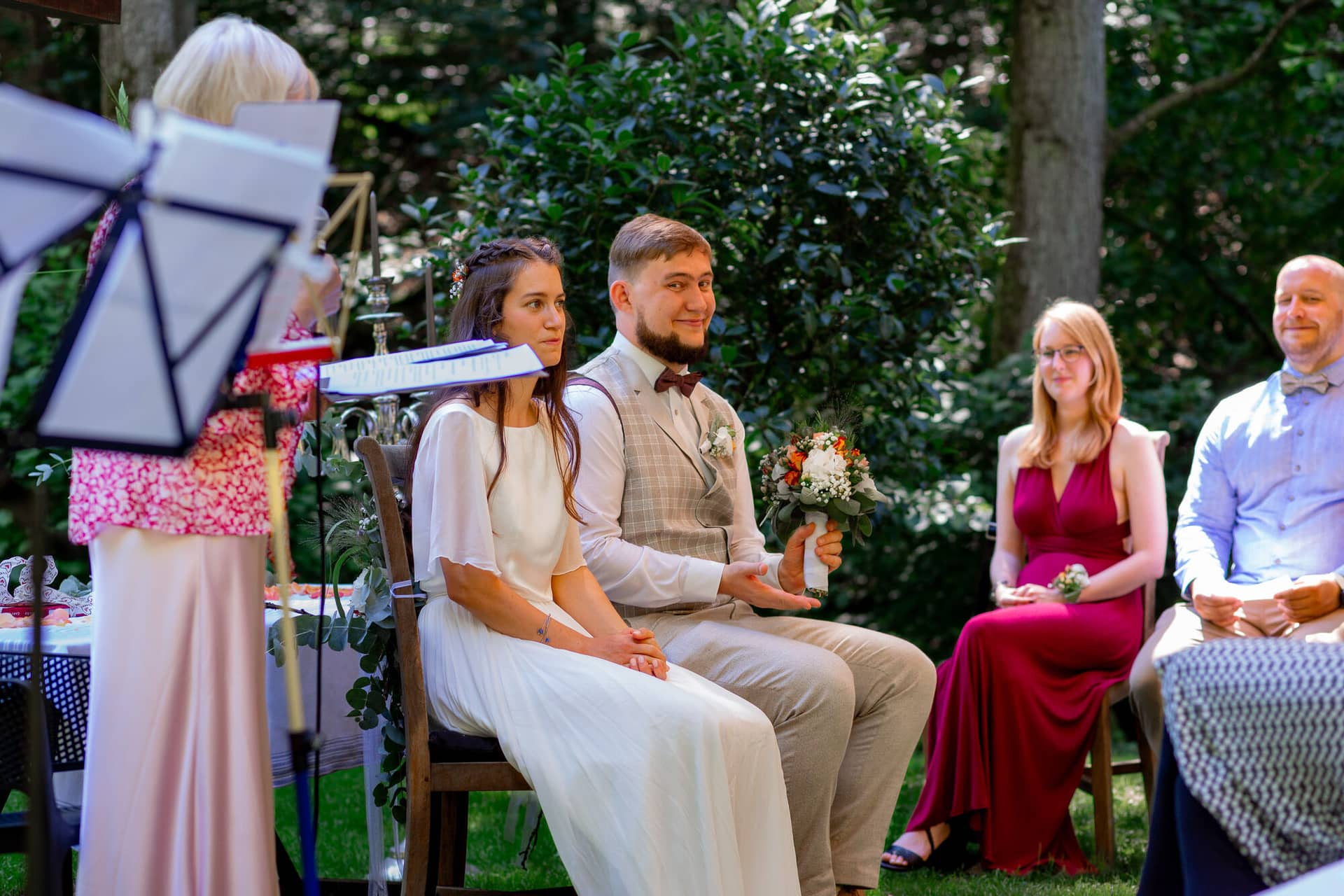 20230811 14 46 29 Hochzeit Elztal Hochzeitsfotograf Heiraten Heidelberg Landgasthof Heidersbacher Muehle MK3 9044.B1OQlwJD Brautpaar auf Stühlen vor dem Gartenaltar, musikalische Begleitung im Hintergrund