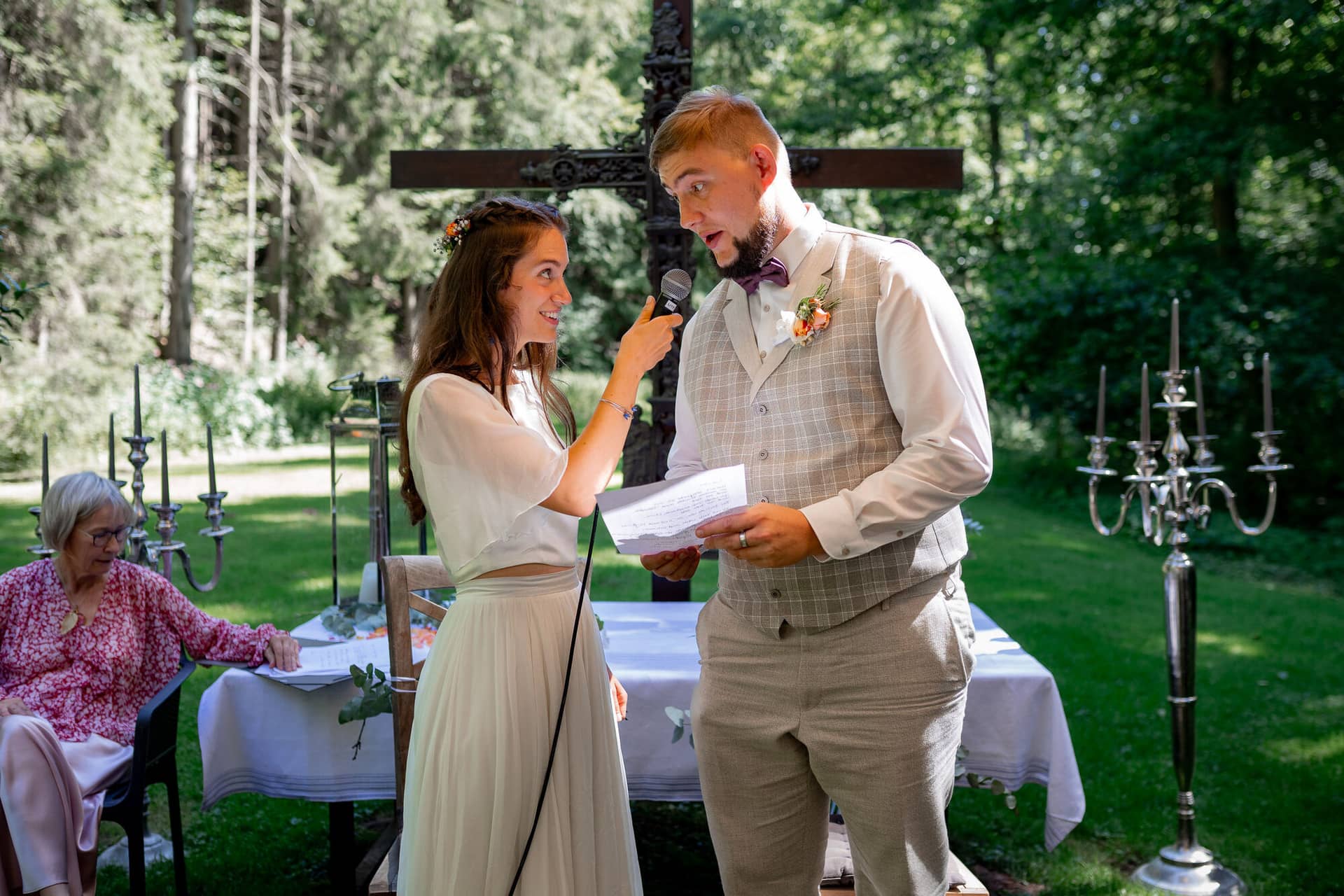 20230811 14 57 42 Hochzeit Elztal Hochzeitsfotograf Heiraten Heidelberg Landgasthof Heidersbacher Muehle 4K8A1514.C04EPWen Bräutigam spricht seine Gelübde mit Mikrofon, Braut reicht ihm lächelnd den Zettel