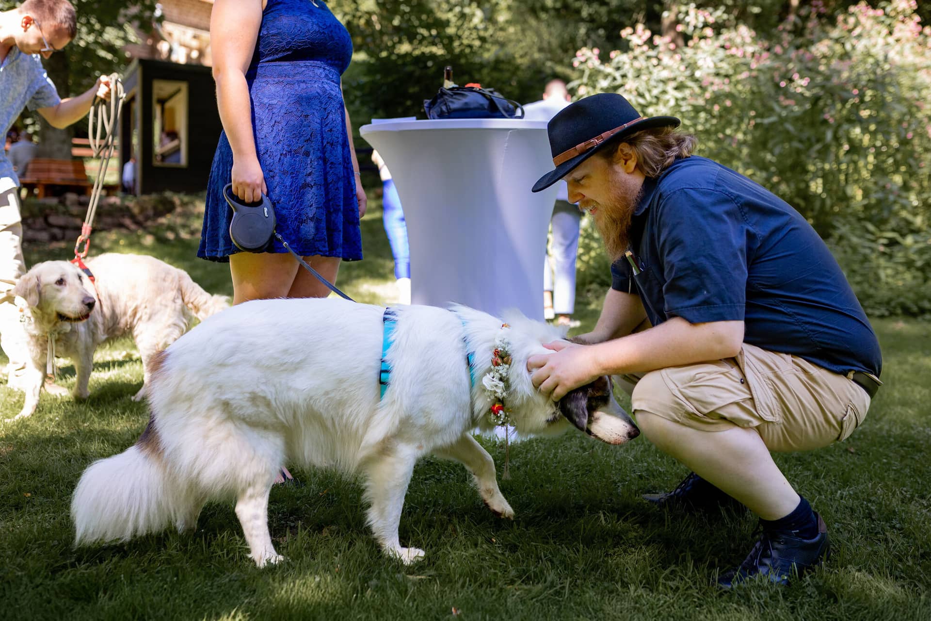 20230811 15 27 08 Hochzeit Elztal Hochzeitsfotograf Heiraten Heidelberg Landgasthof Heidersbacher Muehle 4K8A4270.BUNO3y3e Gast bückt sich hinunter zum weißen Retriever mit Blumenkrone beim fröhlichen Empfang