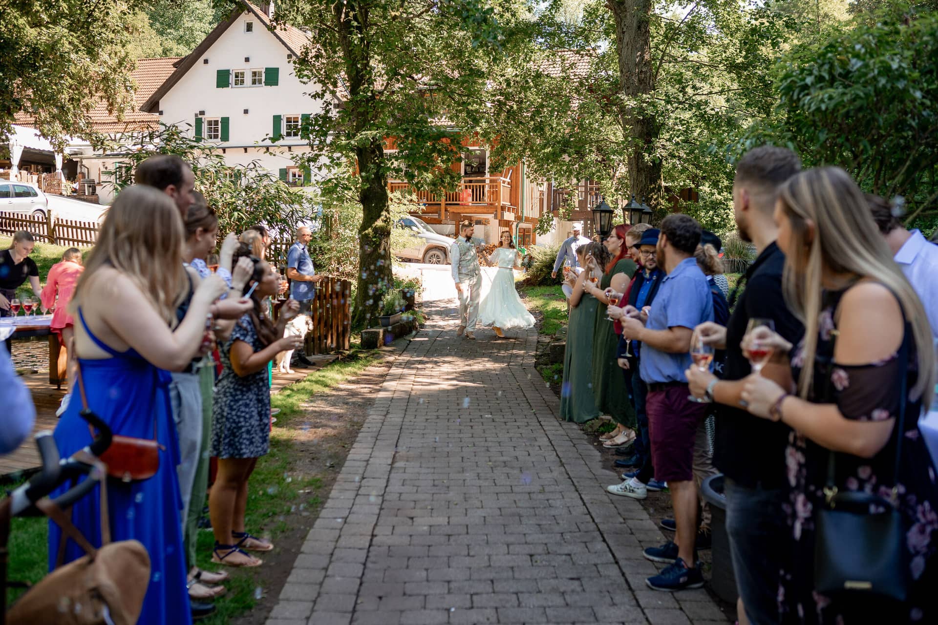 20230811 15 35 17 Hochzeit Elztal Hochzeitsfotograf Heiraten Heidelberg Landgasthof Heidersbacher Muehle 4K8A4576.CBGogs8C Braut geht lächelnd durch die Seifenblasen-Dusche der Gäste zum Holzhaus der Heidersbacher Mühle