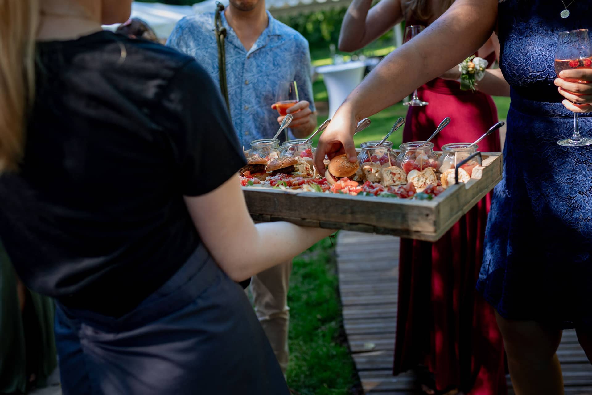 20230811 15 39 31 Hochzeit Elztal Hochzeitsfotograf Heiraten Heidelberg Landgasthof Heidersbacher Muehle 4K8A4951.DsR9PVZ Kellner trägt rustikale Holzplatte mit Vorspeisen und Wein über den sonnigen Rasen