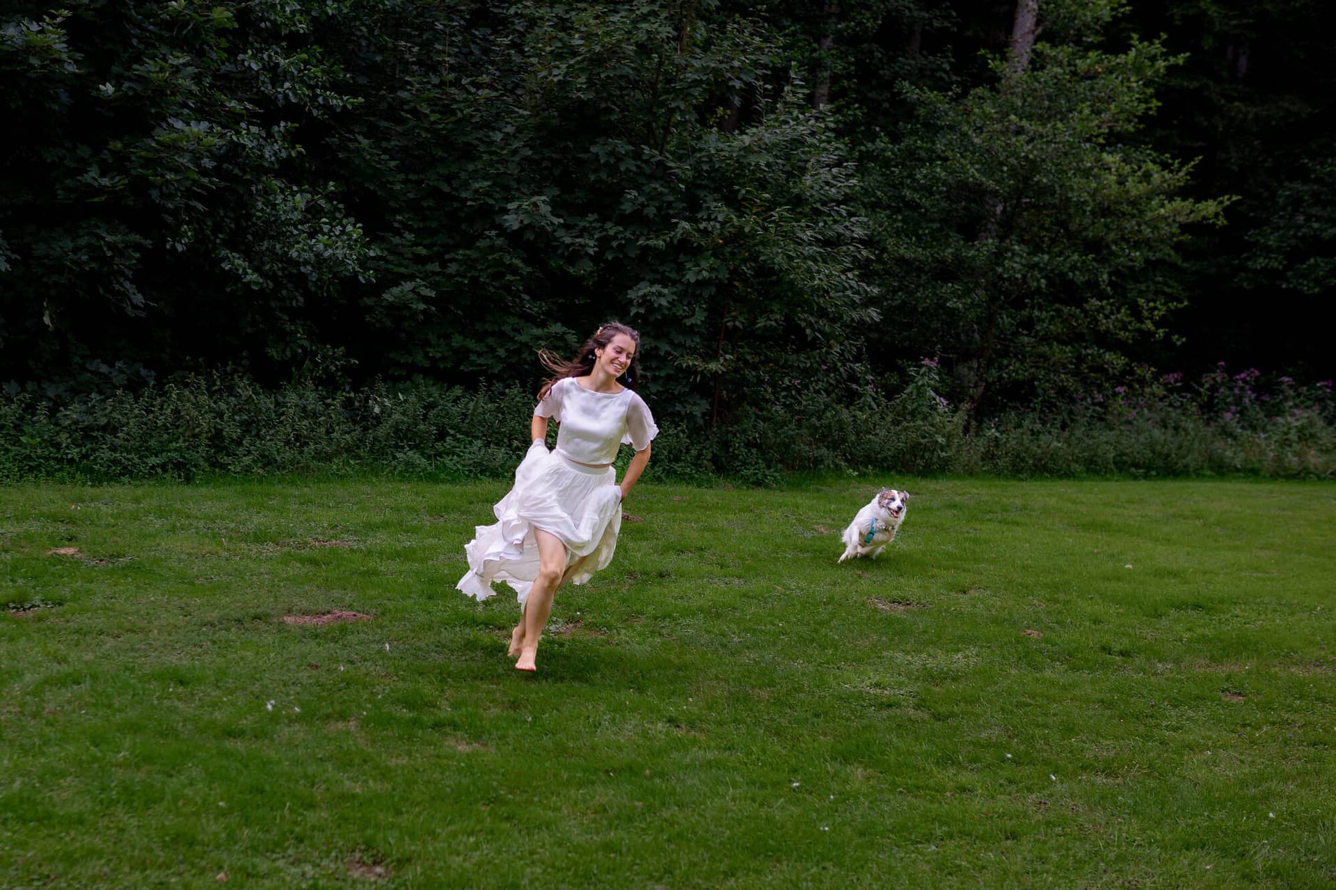 20230811 17 36 23 Hochzeit Elztal Hochzeitsfotograf Heiraten Heidelberg Landgasthof Heidersbacher Muehle 4K8A5911.GFYOFCW1 Braut rennt fröhlich durch die grüne Wiese, Hund folgt begeistert, Elztal-Wald im Hintergrund