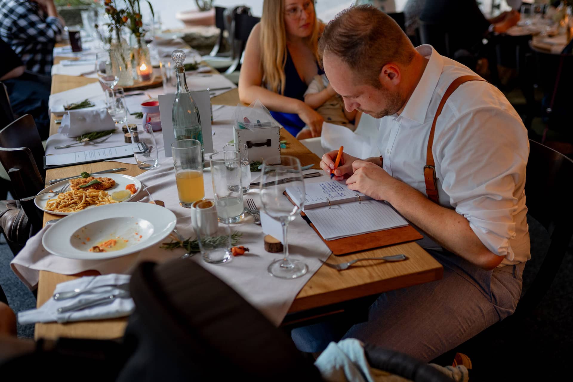 20230811 20 26 12 Hochzeit Elztal Hochzeitsfotograf Heiraten Heidelberg Landgasthof Heidersbacher Muehle 4K8A7651.BAFtMv14 Hochzeitsspiel beim Abendessen: Gast schreibt ins Notizbuch bei Pasta und Wein