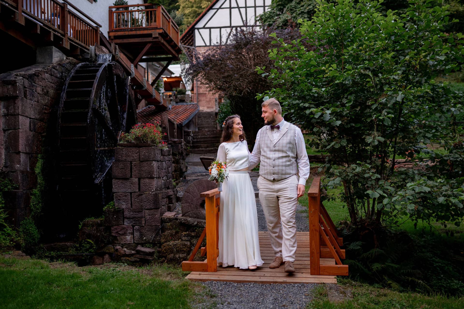 20230811 20 41 27 Hochzeit Elztal Hochzeitsfotograf Heiraten Heidelberg Landgasthof Heidersbacher Muehle 4K8A7851.CIgT8b9C Brautpaar steht auf der kleinen Holzbrücke über den malerischen Wasserfall der Heidersbacher Mühle
