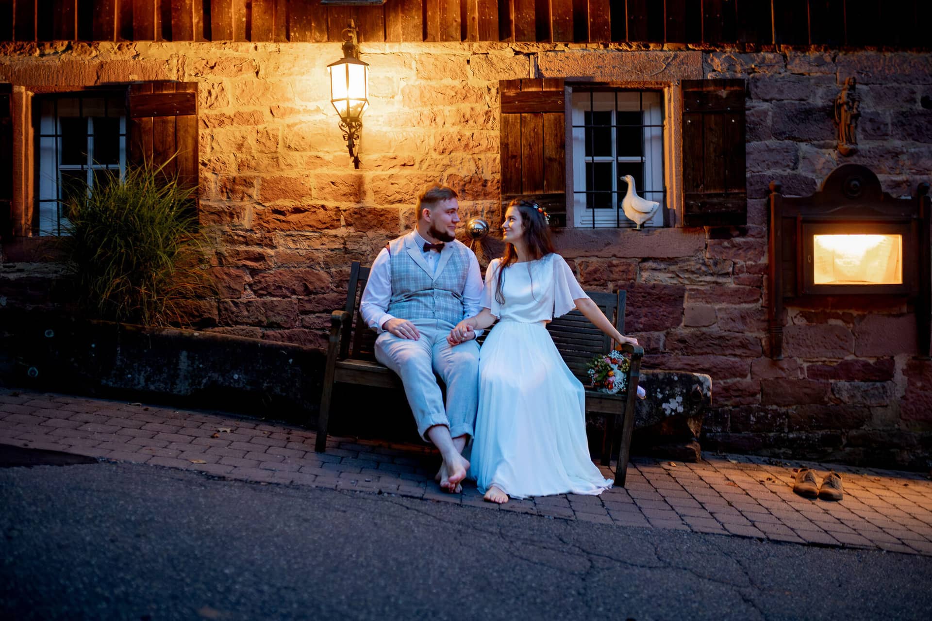20230811 21 13 12 Hochzeit Elztal Hochzeitsfotograf Heiraten Heidelberg Landgasthof Heidersbacher Muehle 4K8A9011 Verbessert RR.BiE IDJg Brautpaar sitzt auf alter Steinbank vor dem Häuschen der Mühle, weiße Ente passiert den Weg