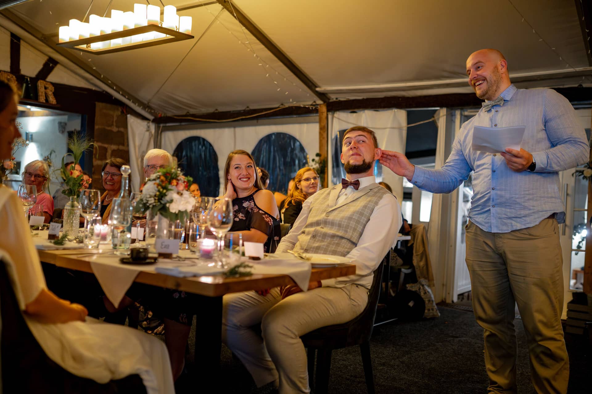 20230811 21 40 44 Hochzeit Elztal Hochzeitsfotograf Heiraten Heidelberg Landgasthof Heidersbacher Muehle 4K8A9135 Verbessert RR.BSpl3jp5 Gast liest unterhaltsamen Text vor, Gäste am Tisch lachen laut beim Hochzeitsspiel