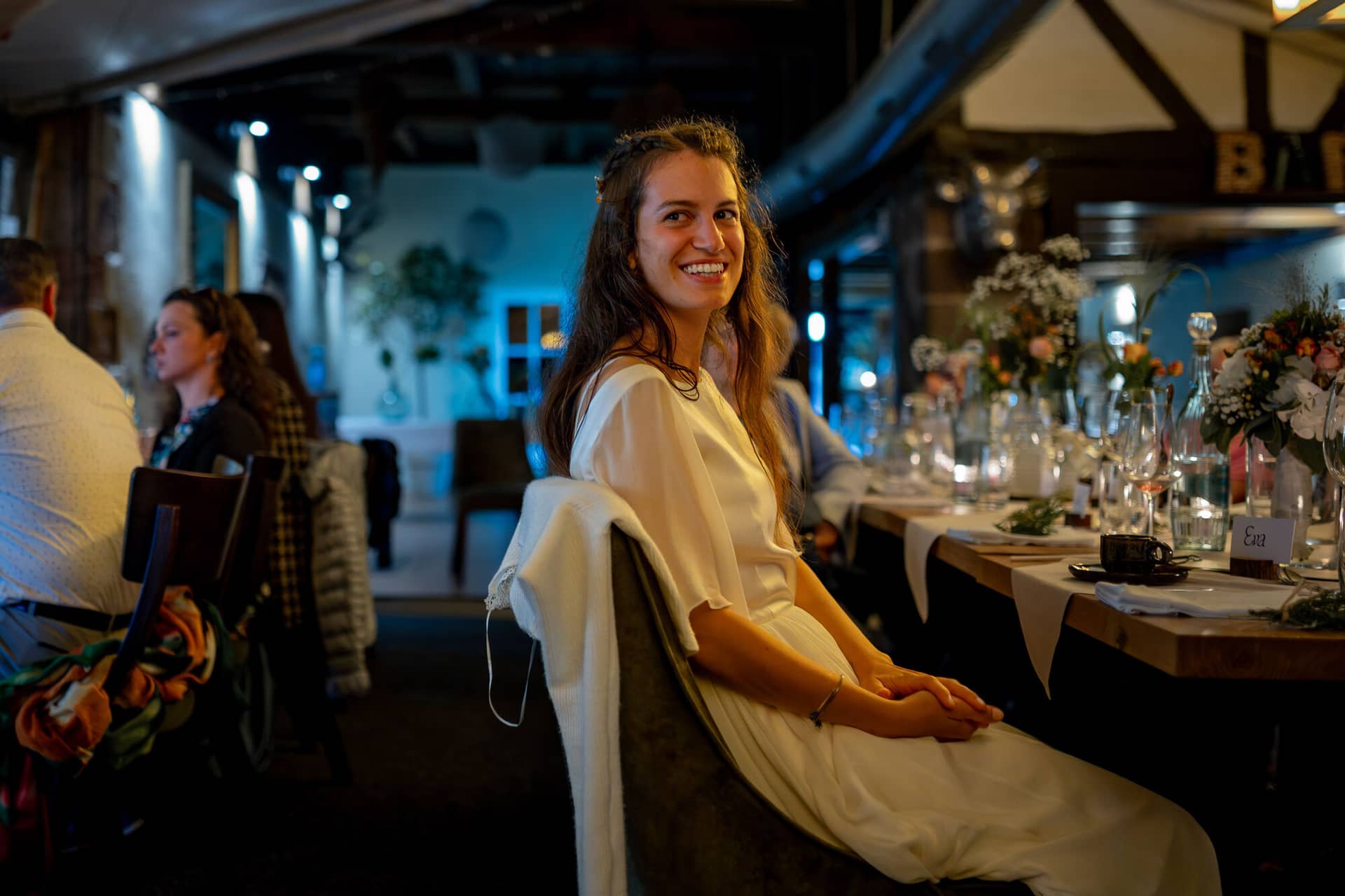 20230811 21 41 32 Hochzeit Elztal Hochzeitsfotograf Heiraten Heidelberg Landgasthof Heidersbacher Muehle 4K8A9189 Verbessert RR.BNSTdT2O Braut sitzt entspannt am Abendtisch und genießt den Moment, Gäste feiern im Hintergrund