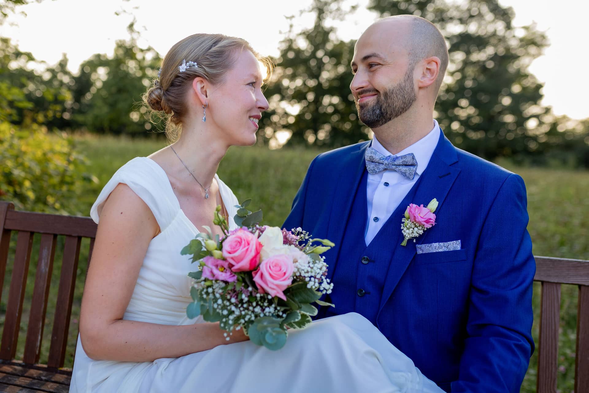 20240820 19 12 27 Teusch Schlossgarten Worms Herrnsheim 4K8A7590.CFCWmEii Brautpaar sitzt lächelnd auf einer Holzbank auf einer Wiese im Schlossgarten Herrnsheim