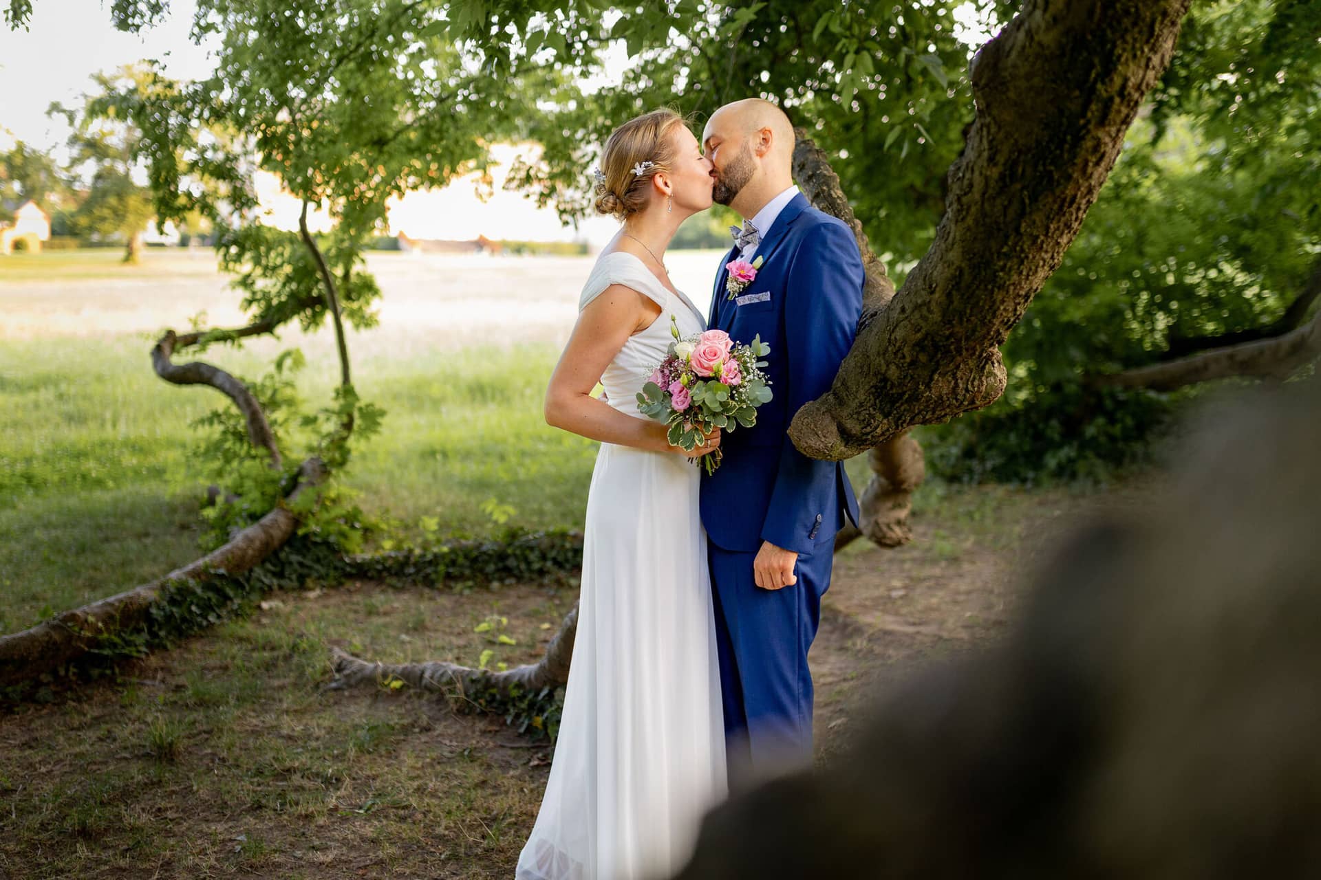 20240820 19 18 47 Teusch Schlossgarten Worms Herrnsheim 4K8A7872 Hochzeitsfotograf Matthias Butz.Y8lvYQUR Brautpaar küsst sich unter einem großen knorrigen Baum auf einer Wiese im Schlossgarten