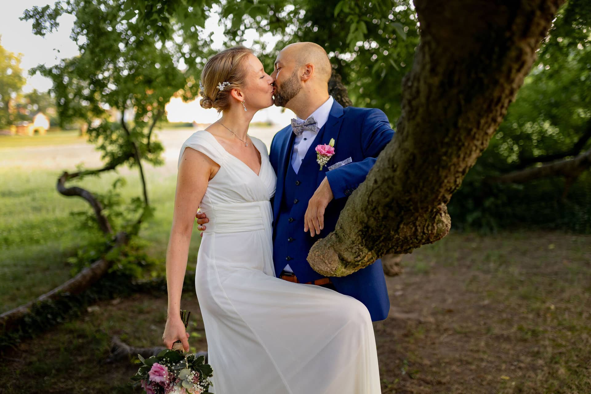 20240820 19 19 36 Teusch Schlossgarten Worms Herrnsheim 4K8A7959 Hochzeitsfotograf Matthias Butz.KOR9KPla Brautpaar küsst sich unter den Ästen eines alten Baumes auf grüner Wiese im Schlossgarten Herrnsheim
