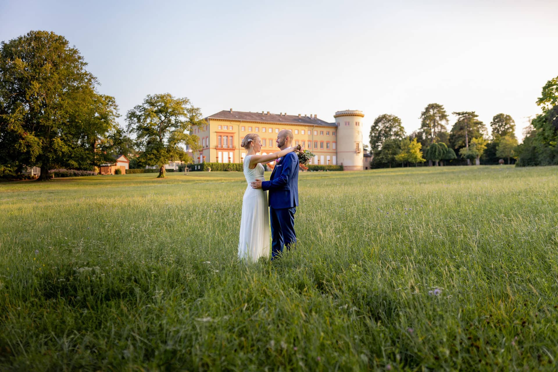 20240820 19 22 26 Teusch Schlossgarten Worms Herrnsheim 4K8A8001 Hochzeitsfotograf Matthias Butz.Y3U74DJk Brautpaar umarmt sich eng in hohem grünen Gras, im Hintergrund das gelbe Schloss Herrnsheim