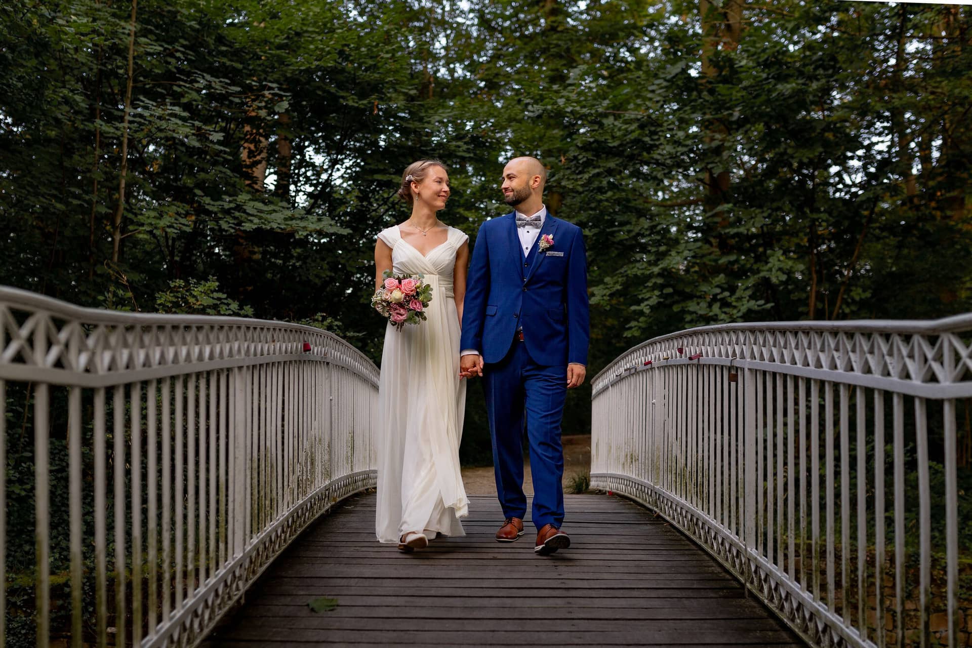20240820 19 40 35 Teusch Schlossgarten Worms Herrnsheim 4K8A8895.BomgqfaR Brautpaar geht Hand in Hand über eine Holzbrücke durch einen dichten Waldbereich