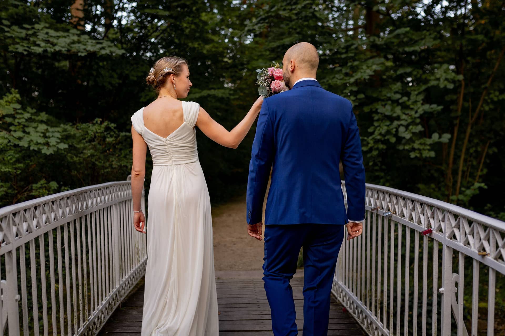 20240820 19 42 08 Teusch Schlossgarten Worms Herrnsheim 4K8A8970.B5A8DWr2 Brautpaar geht Hand in Hand über die Holzbrücke im Wald, Braut im weißen Kleid, Bräutigam im dunkelblauen Anzug