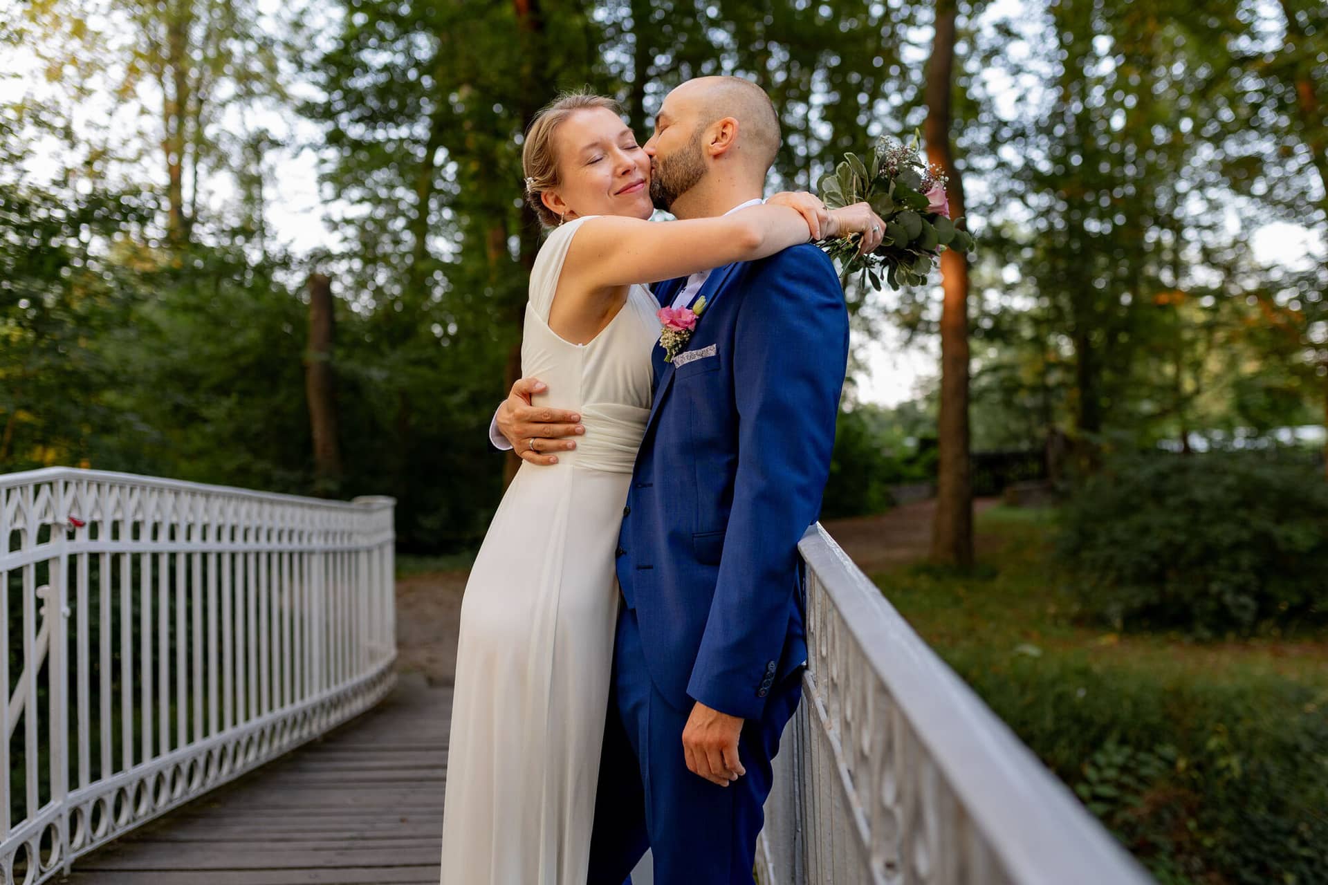 20240820 19 47 02 Teusch Schlossgarten Worms Herrnsheim 4K8A9197 Hochzeitsfotograf Matthias Butz.CjekG6DD Bräutigam umarmt Braut auf der Holzbrücke im Wald, Braut hält kleinen Blumenstrauß