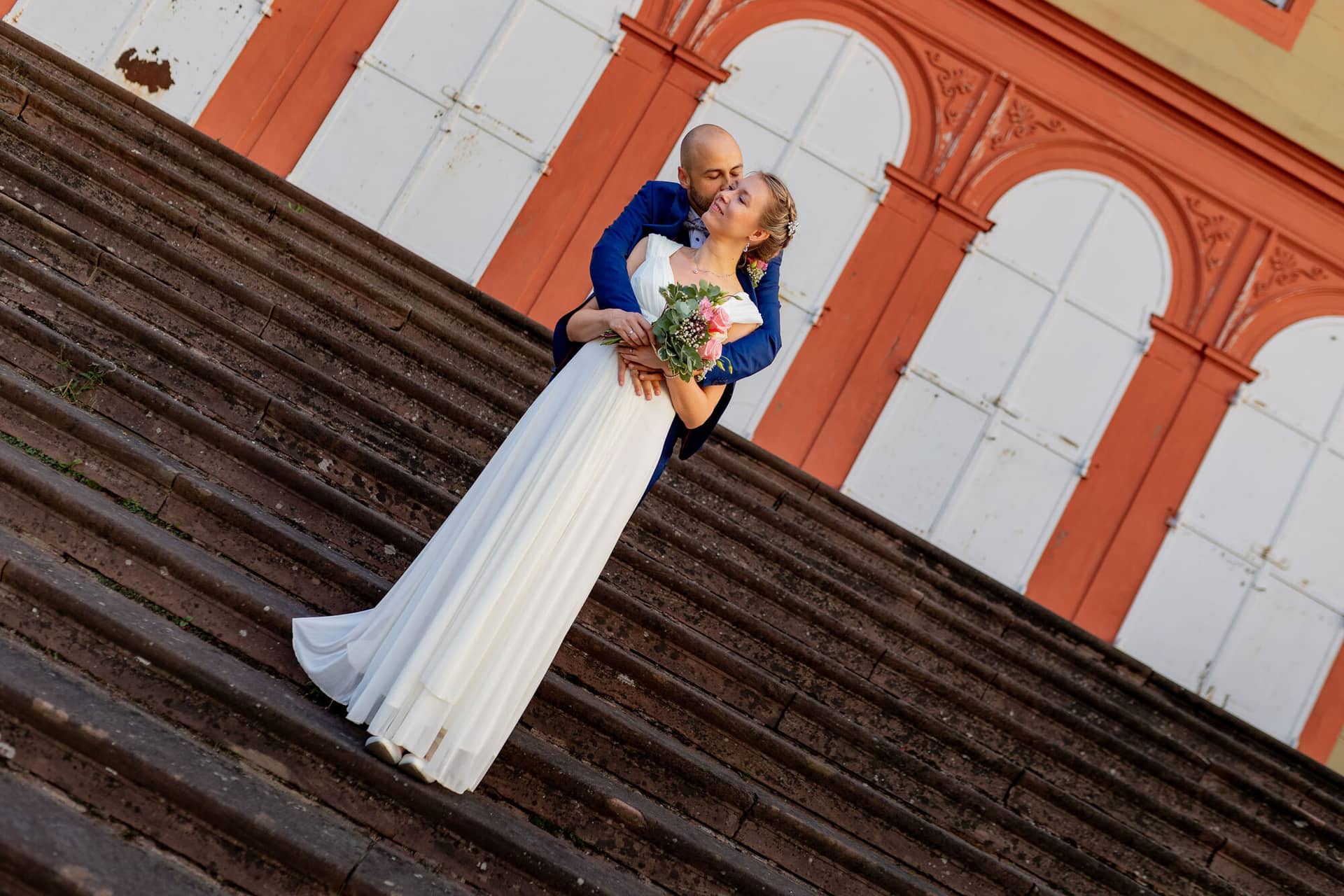 20240820 20 00 34 Teusch Schlossgarten Worms Herrnsheim 4K8A9814 Hochzeitsfotograf Matthias Butz.CZqrHabw Brautpaar küsst sich auf den Stufen eines historischen Backsteingebäudes im Schlossgarten