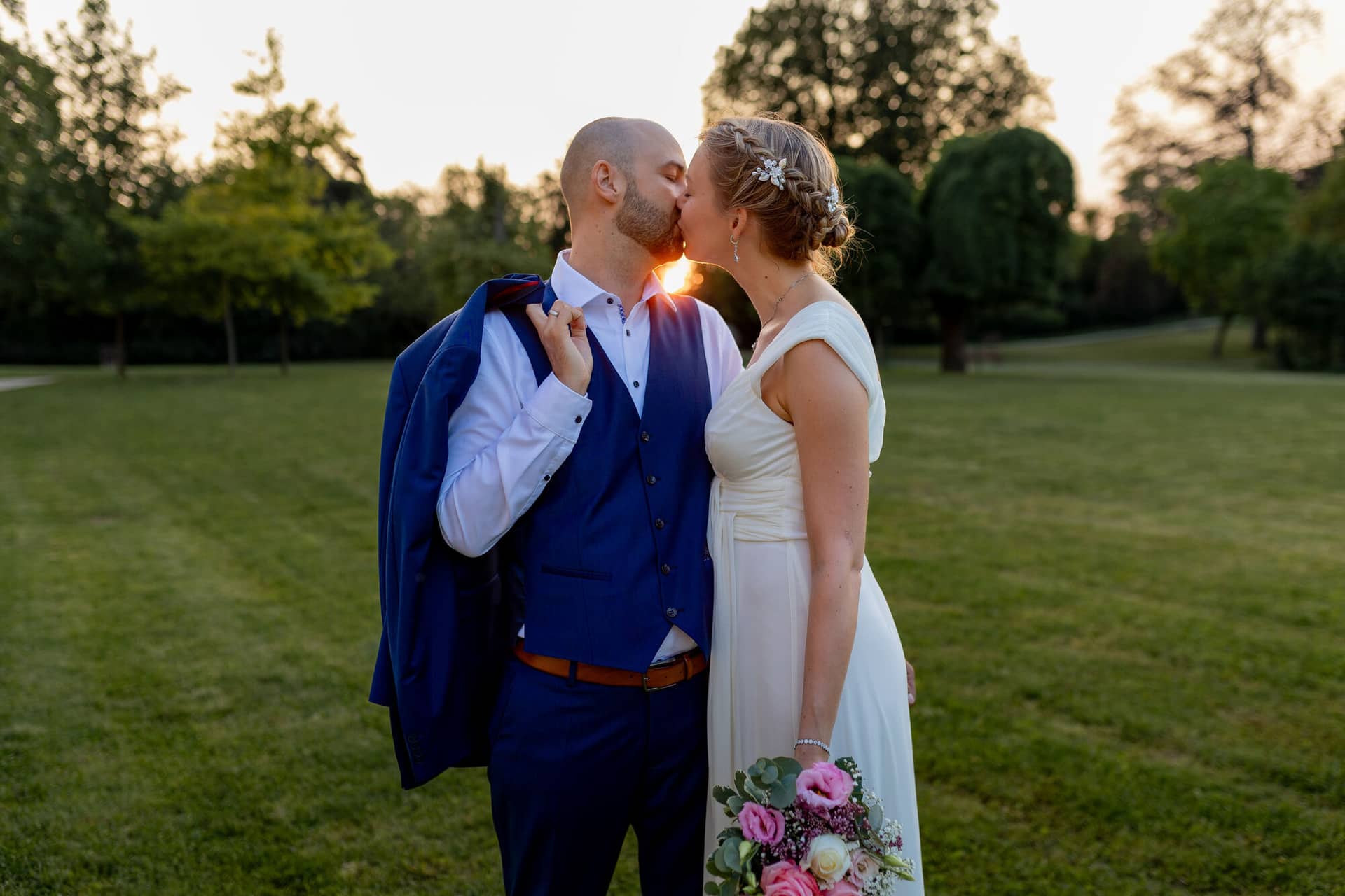 20240820 20 05 56 Teusch Schlossgarten Worms Herrnsheim 4K8A0136.BnyHaoJY Brautpaar küsst sich auf einer Wiese im goldenen Sonnenuntergangslicht des Schlossgartens Herrnsheim